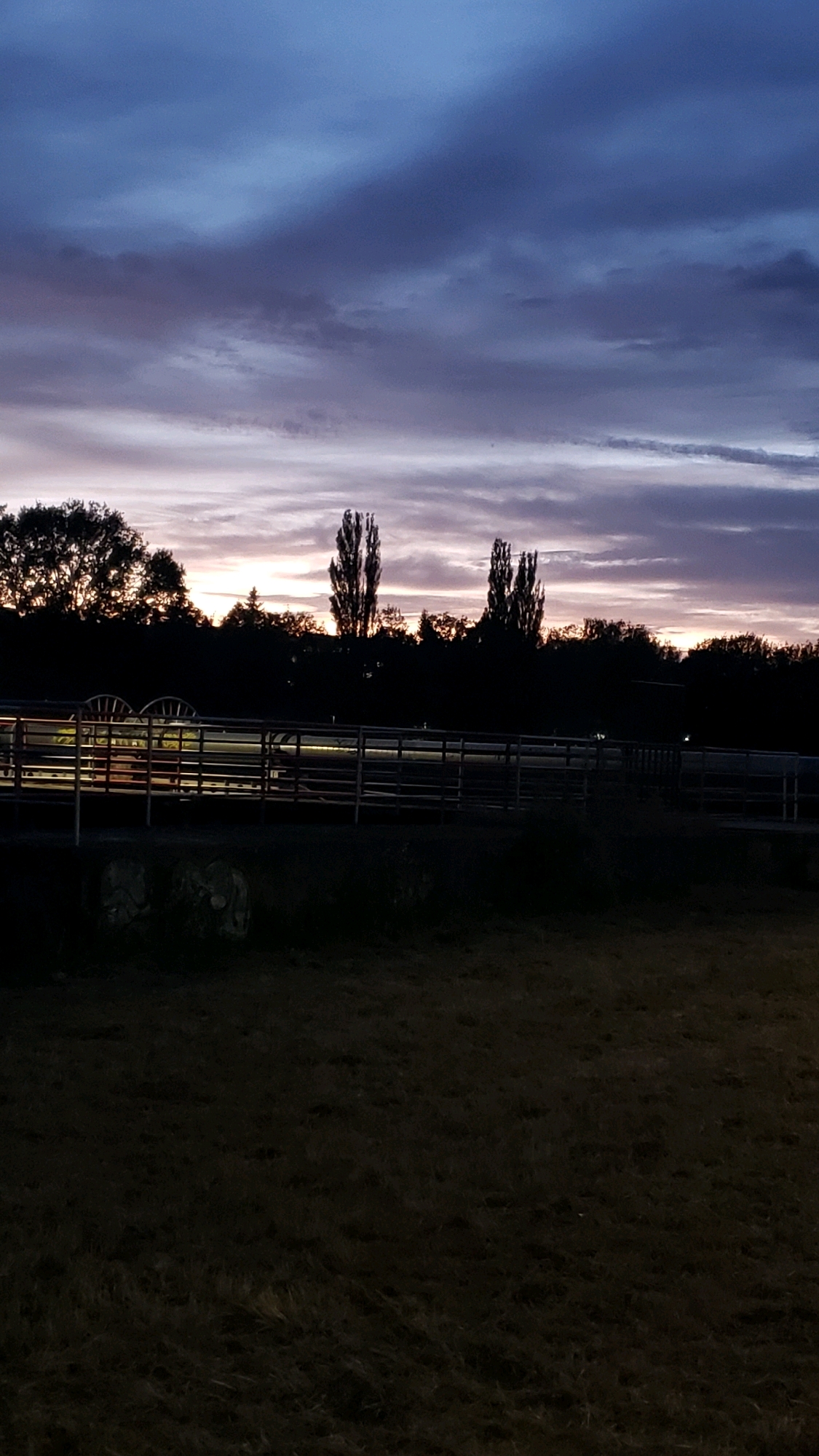The image depicts a rural scene at dusk, with a dramatic sky featuring shades of dark blue, purple, and orange. Silhouetted against the sky are trees and tall, slender poplar trees. In the middle ground, a fence with horizontal rails runs across the frame, with some circular objects, possibly hay bales, visible behind it. The foreground is a dark, grassy field. The scene is likely located outside of the city of Prešov, Slovakia, given the rural characteristics. No people or specific actions are discernible.