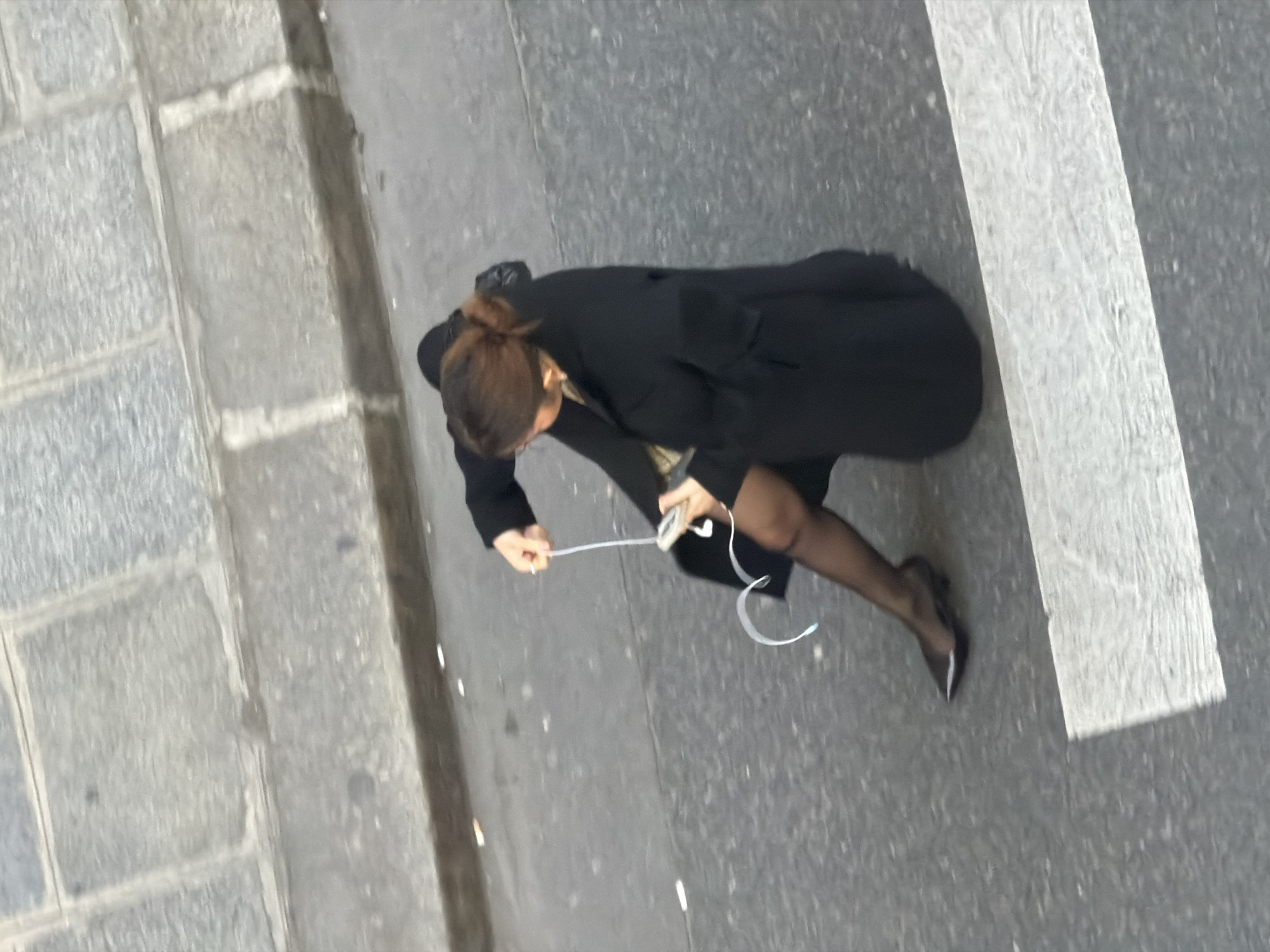 The image shows a woman from an aerial perspective, standing on a city street in what appears to be Paris, France. The city street has sections of grey brick and a white pedestrian crossing. She is wearing black stockings, black heels, and a black coat. She has brown hair pulled up in a bun. She is holding a mobile phone and tangled headphones in her hands.
