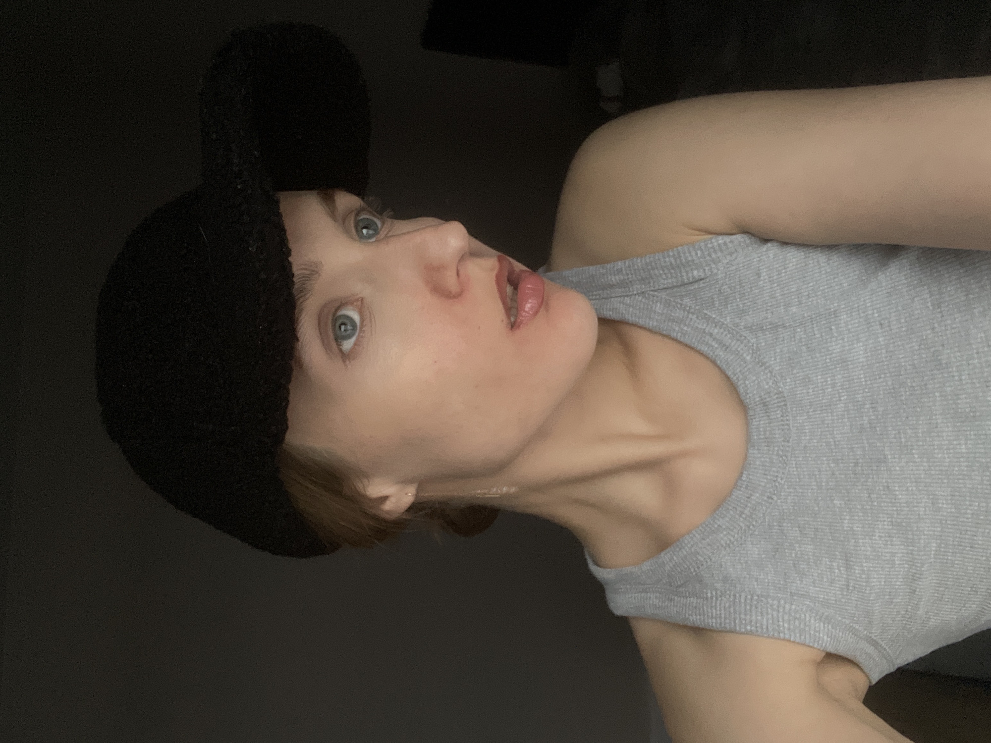 A woman with fair skin and light blue eyes is pictured wearing a gray tank top and a black cap. She is looking up and to the left. The background is dark and out of focus. There is no indication in the image that the location is Подшивалово, Russia.