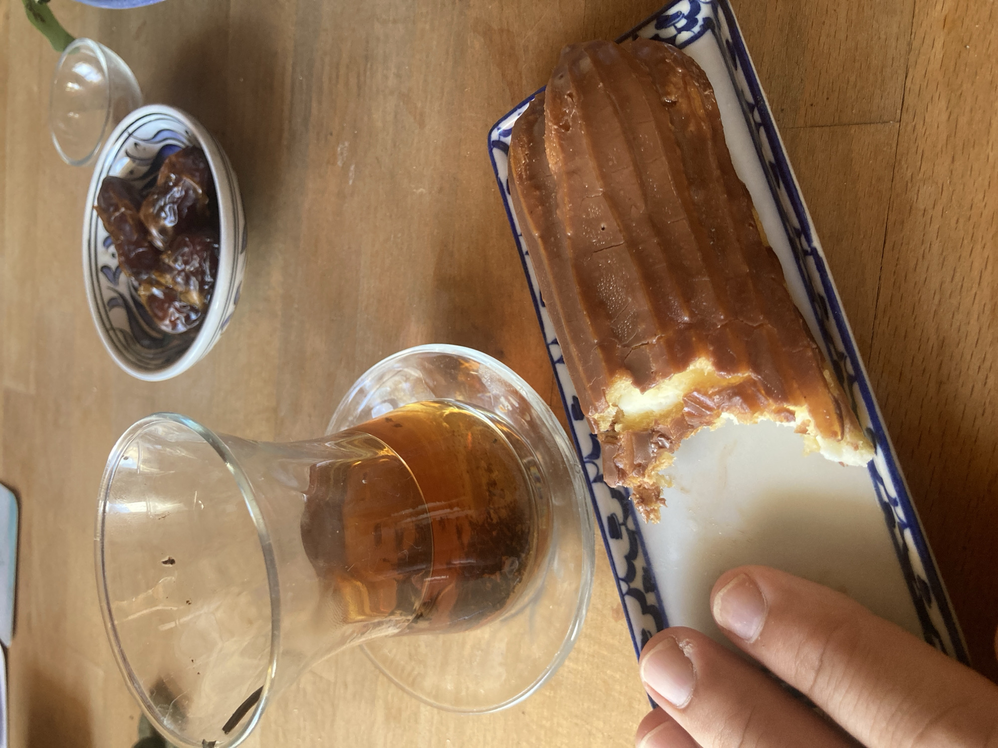 A close-up, top-down shot showcases a table setting with a partially eaten éclair, a glass of amber-colored liquid, and a small bowl of dates. The éclair, which appears to be filled with cream and topped with a chocolate glaze, has a bite taken out of it. It rests on a rectangular white plate with a blue and white patterned rim. Beside the éclair, a clear glass with a thick base holds a beverage, possibly tea or coffee, with some dark specks visible at the bottom. To the left of the glass, a small, decorative white bowl is filled with several dates. A tiny, clear glass, possibly a shot glass or a lid, sits next to the bowl. A person's hand, with visible fingernails, is positioned in the lower right corner of the frame, reaching towards the éclair. The table surface appears to be made of wood. The lighting suggests it could be daytime, possibly indoors with natural light. The scene evokes a sense of a relaxed break or a casual snack. There is no visible text in the image.