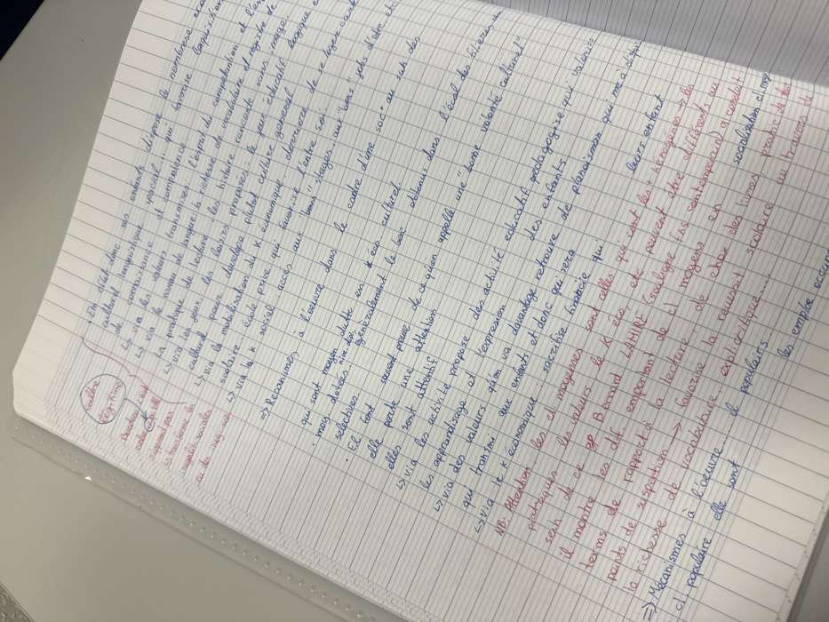 The image displays a close-up, top-down view of an open, spiral-bound notebook. The paper features French-style Séyès ruling, characterized by horizontal blue lines with faint vertical red lines forming a grid. The notebook rests on a light-colored, possibly white or off-white, flat surface. A translucent, corrugated plastic sheet, typical of a folder protector or binder, is visible along the left edge of the notebook.
The pages are filled with extensive handwritten notes primarily in blue ink, with certain words, phrases, and annotations highlighted or written in red ink. The text is in French, with structured bullet points, arrows, and circled terms. Key subjects mentioned in the notes include "culture," "régime," "inégalité sociales" (social inequalities), "Bourdieu," "Bernard Lahire," "capital économique" (economic capital), and "capital culturel" (cultural capital), indicating a focus on sociology or education. One section in red ink is circled and labeled "culture" and "régime," with arrows connecting to related concepts. No individuals are present in the scene, and no specific city location, such as Albi, France, is discernible from the visual information provided within the image frame.