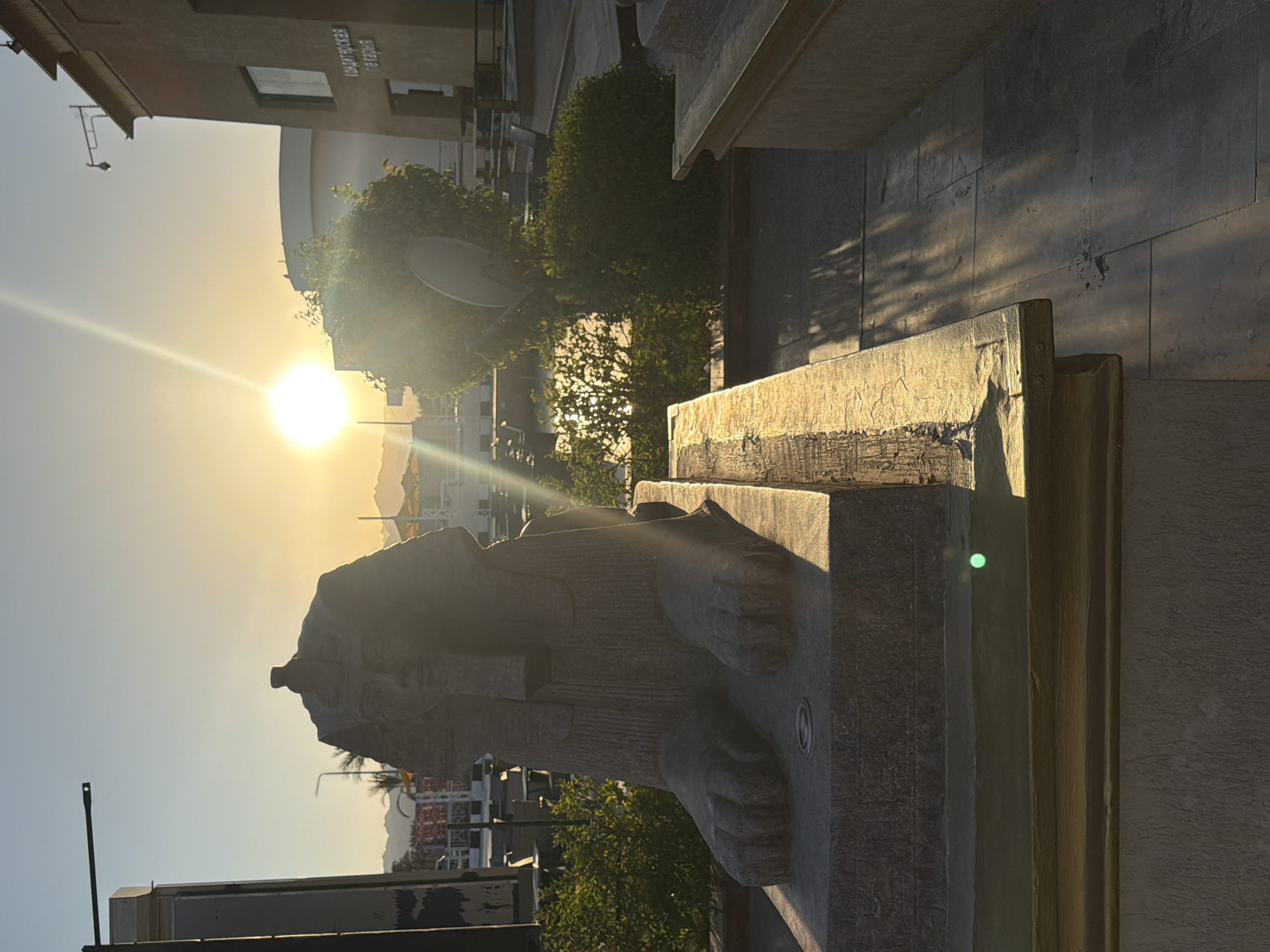 The image captures a sphinx statue in Sharm El Sheikh, Egypt, during sunset. The sun, a bright orb in the sky, casts long shadows and illuminates the scene with a warm, golden light. The statue, made of stone, is intricately carved and detailed, showing its lion body and human head. It appears to be a decorative element in a public space, possibly a park or a promenade, given the presence of buildings and trees in the background.
The setting suggests an outdoor environment, with a clear sky and the characteristic warm hues of a desert sunset. The surrounding architecture is modern, with clean lines and light-colored facades. Lush green trees provide a contrast to the stone of the statue and the buildings. The overall mood is serene and peaceful, with the setting sun creating a tranquil atmosphere. There is no visible text in the image. The orientation of the image is portrait, with the sky and buildings appearing on the left side and the statue and its pedestal dominating the right.