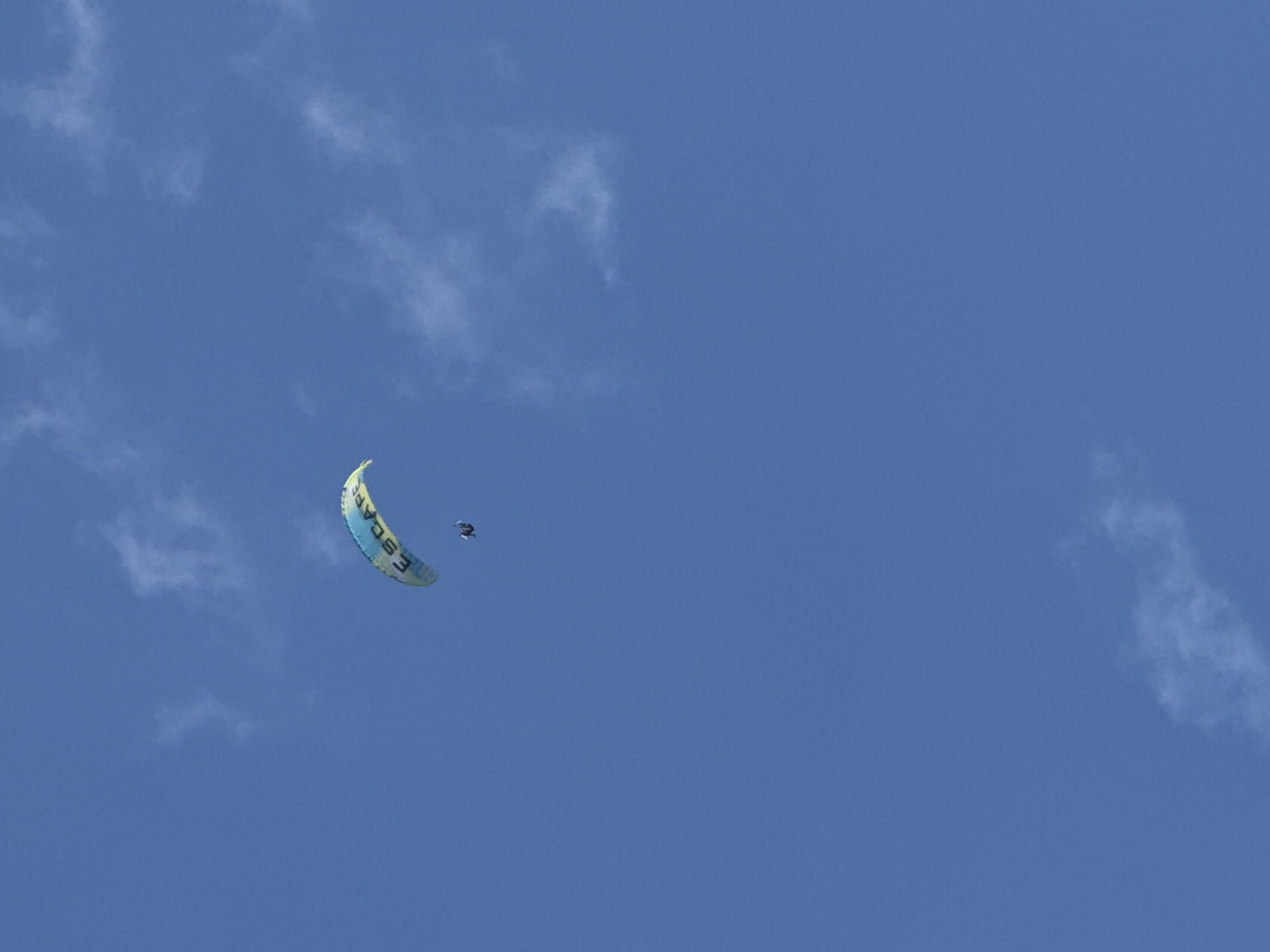 A paraglider is captured mid-flight against a clear blue sky with scattered white clouds. The paraglider is yellow and blue with the word "ESCAPE" printed on it. A person is attached to the paraglider, appearing small against the vast expanse of the sky. The image is taken from below, looking upwards at the paraglider. This activity is common in Fethiye, Turkey, particularly from the famous Babadağ mountain, known for its paragliding launch sites.