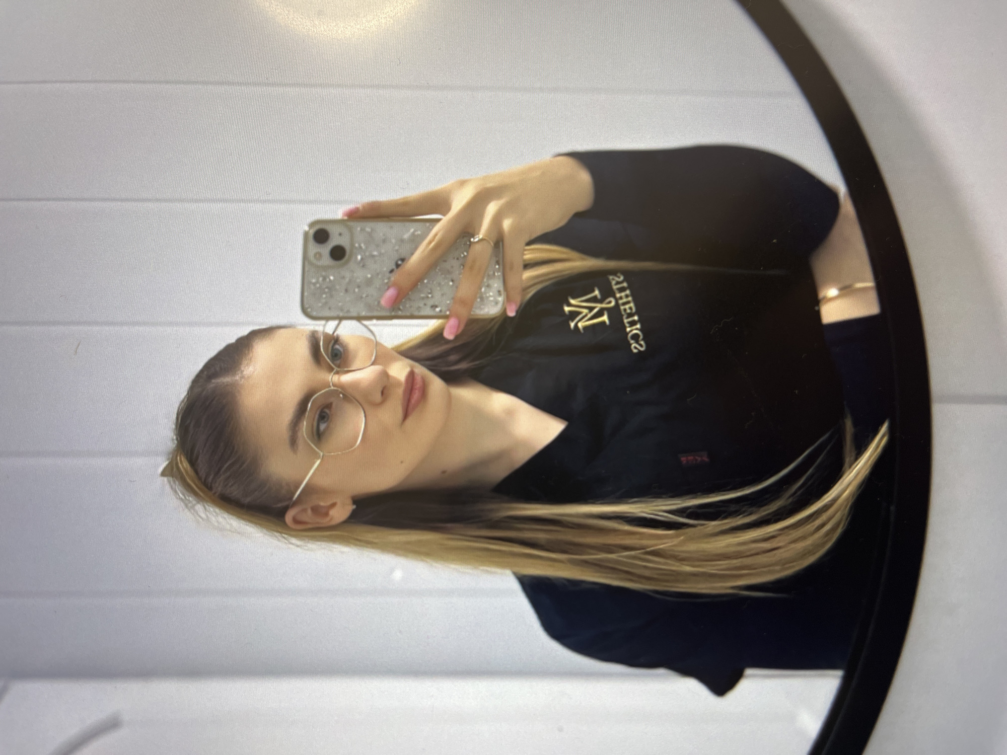 A young woman is seen in a mirror taking a selfie with her cell phone. The woman has long blonde hair and is wearing glasses and a dark-colored shirt with the word "ATHLETICS" printed on it. She has on a ring on her finger and a bracelet on her wrist. The background of the image includes white horizontal lines. The location is Unknown, Unknown.