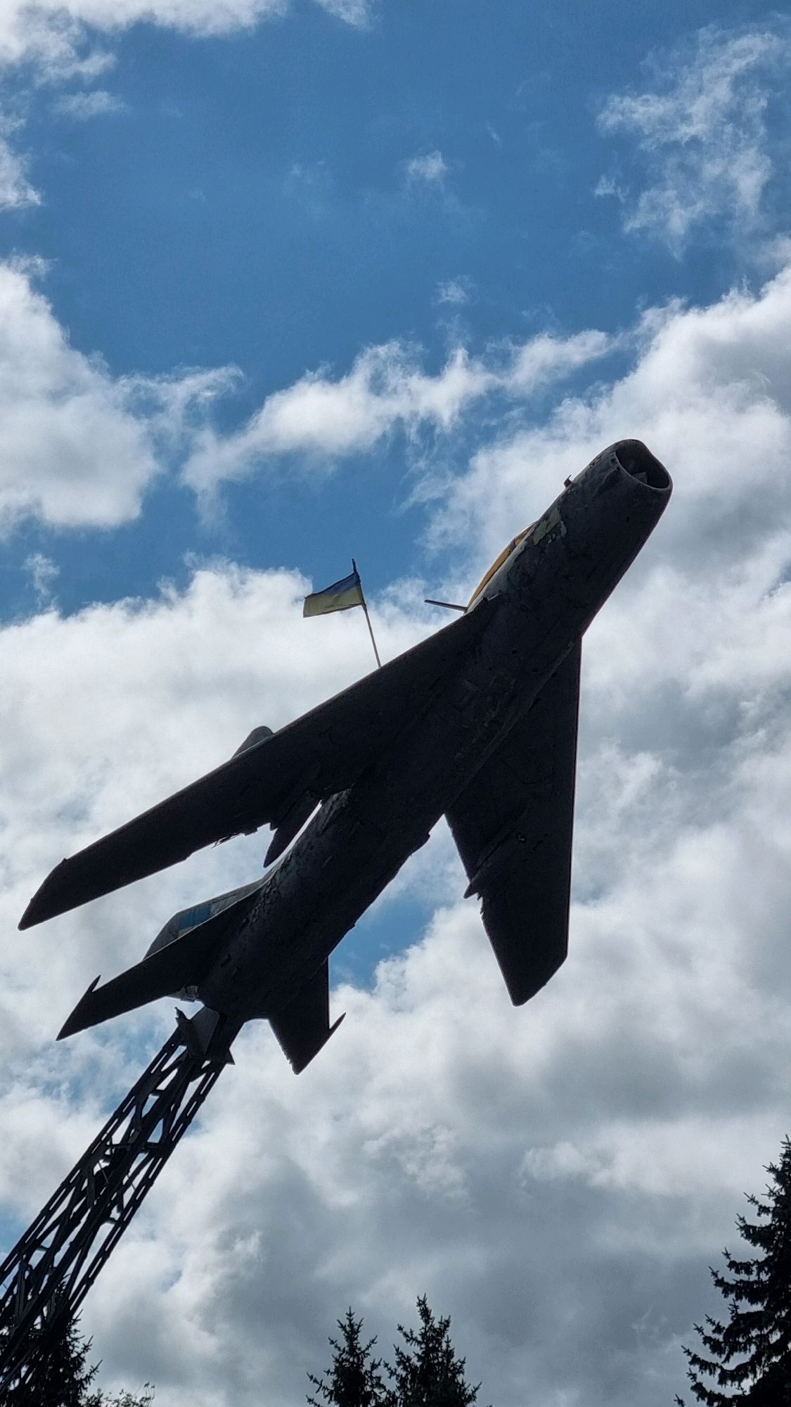 An upward, low-angle shot captures a dark silhouette of a monument to a Soviet-era jet fighter against a sky filled with white, cumulus clouds. The fighter jet is mounted on a tall, dark metal pylon, angled upwards and to the left. A small Ukrainian flag, yellow and blue, is attached to a pole on the jet's wing. The jet's nose cone is pointed towards the top right of the frame, and its wings are extended. The sky is a bright blue, with patches of sunlight breaking through the clouds. In the bottom corners of the image, dark green, pointed evergreen trees are visible, suggesting an outdoor, possibly park-like setting. The overall impression is one of a memorial or monument in an open space. The specific location is a monument to a MiG-17 fighter jet, a memorial to Ukrainian Air Force pilots, located in the city of Dubno, Ukraine.