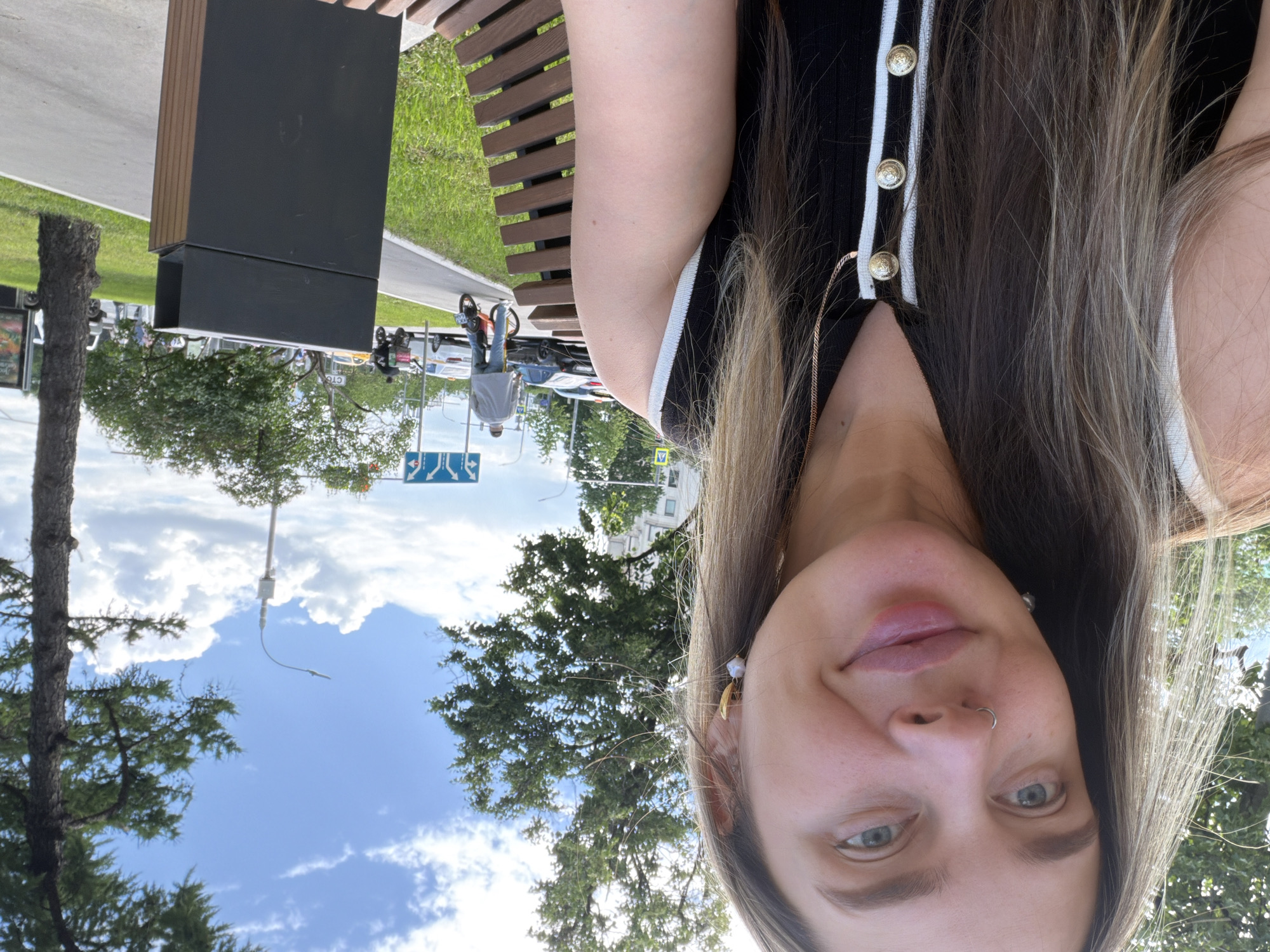 The image is a selfie taken by a woman with light skin, light brown hair, and light eyes. She wears a black and white striped top with gold buttons. She has a nose ring and a gold earring. The background reveals an outdoor scene in what appears to be a city park. There is a wooden bench beside her. In the background, there's a road with cars, trees, a black modern structure (perhaps a bench or trash receptacle), and a sign indicating road directions. The sky is visible with scattered clouds. The image seems to be taken in Podolsk, Russia, based on the road sign.