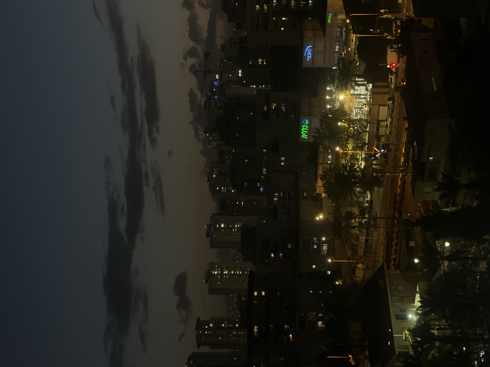 The image shows a nighttime view of Kiryat Bialik, Israel. The city's skyline is visible with lit buildings against a dark sky with clouds. On the right side, a lit area includes roads, palm trees, and buildings with signs in Hebrew, including "Clalit" and "Maccabi". Traffic lights are also visible. The image suggests an urban environment at dusk or night.
