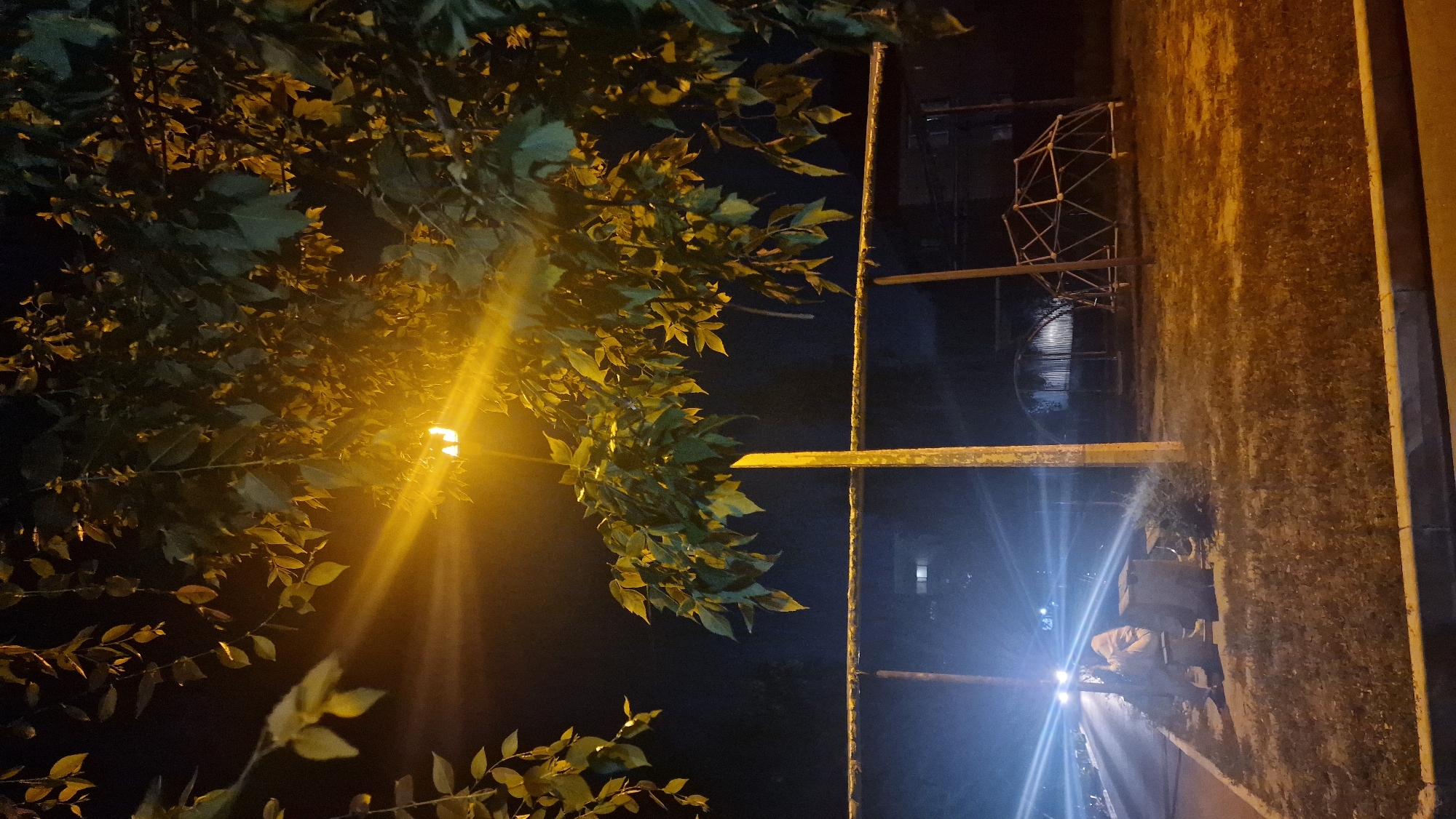 The image captures an outdoor nighttime scene. A bright street lamp, with its light rays radiating outwards, illuminates a tree with green leaves. In the foreground, a grassy area with a curb is visible. Further back, a metal railing runs horizontally across the frame. To the left, a person is sitting on a bench near a trash receptacle, with their back to the viewer. The bright headlights of an approaching vehicle are visible on a road in the background. To the right, a geometric playground structure stands in the dark. The overall environment suggests a park or a public recreational area.