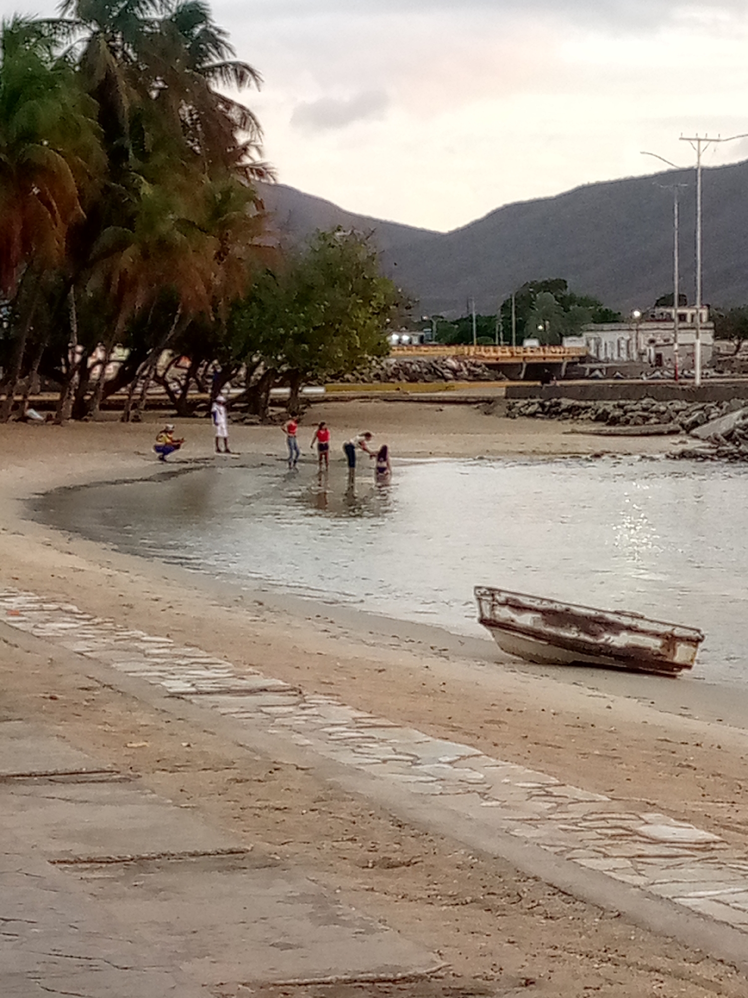 This image captures a tranquil coastal or lakeside scene, likely during dusk or late afternoon, under an overcast sky. The foreground features a sandy beach with a textured, possibly tiled or cobbled, pathway running along its edge from the lower left to the middle right. A small, weathered, light-colored boat is beached on the sand to the right.
In the mid-ground, several individuals are gathered near and in the calm, shallow water. On the sandy shore, one person in a white top stands by the water's edge, while another, wearing a yellow top, squats nearby. Further into the water, three figures are wading, with one person partially submerged. The activity appears leisurely, suggesting people enjoying the water.
The background is dominated by lush greenery, including numerous palm trees lining the left side of the shore, and other leafy trees. Further back, a yellow or orange bridge spans an inlet or waterway, with buildings visible beyond it. Some of these structures appear to be older, featuring white facades and arched openings. Utility poles with overhead wires are also noticeable near the buildings and bridge. Imposing mountains rise steeply in the distance, forming a dramatic backdrop under the pale sky. The water reflects the soft light, indicating the sun is low or diffused by clouds. No discernible text is visible in the image.