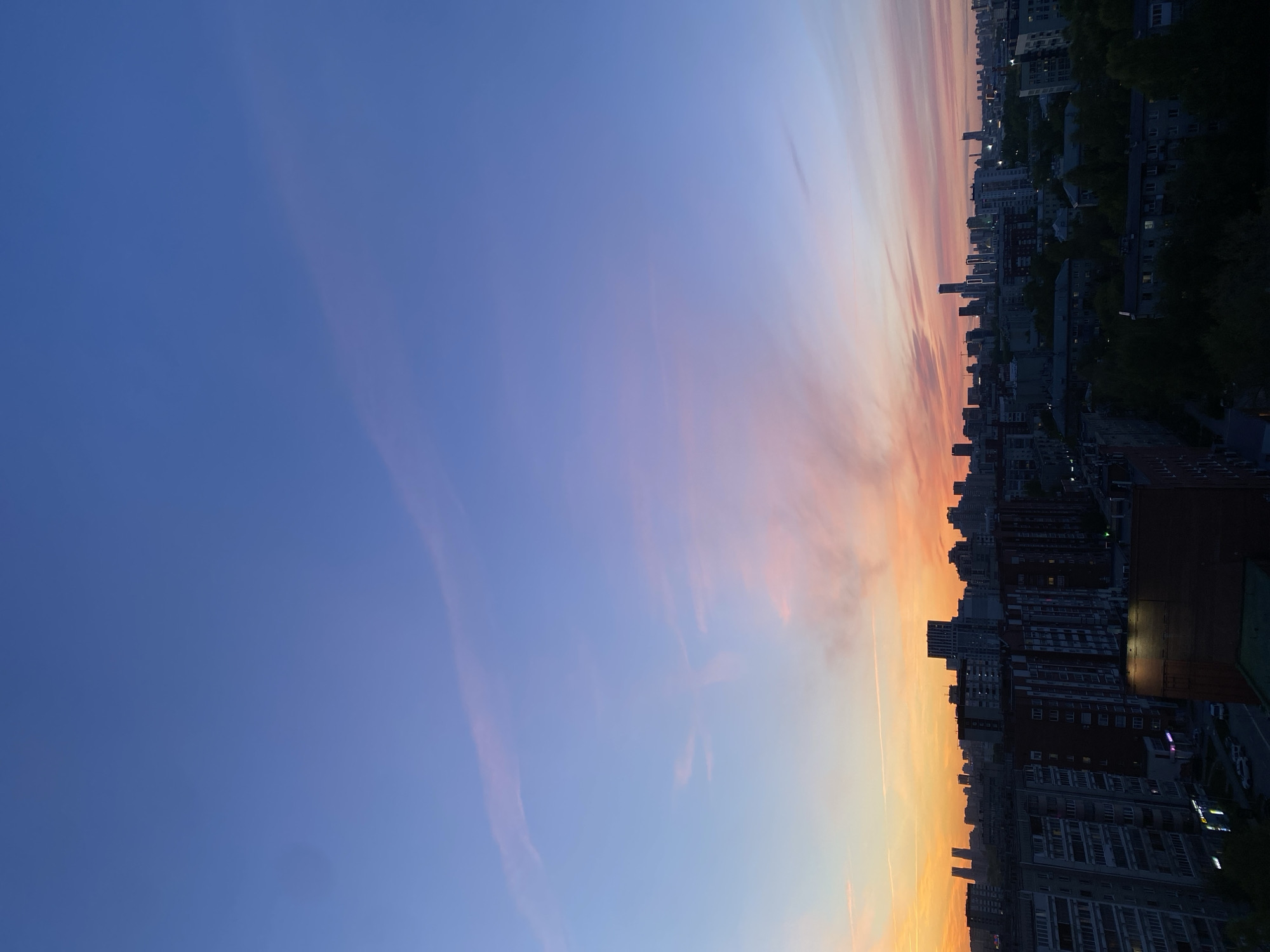 The image captures a sunset over a city, likely Yekaterinburg, Russia, given the architectural style of the buildings. The sky is predominantly blue, transitioning to orange and pink hues towards the horizon. Buildings of various heights and designs form the cityscape, with some trees visible. It appears to be early evening, with the sun setting behind the buildings. No people are directly visible in the frame.