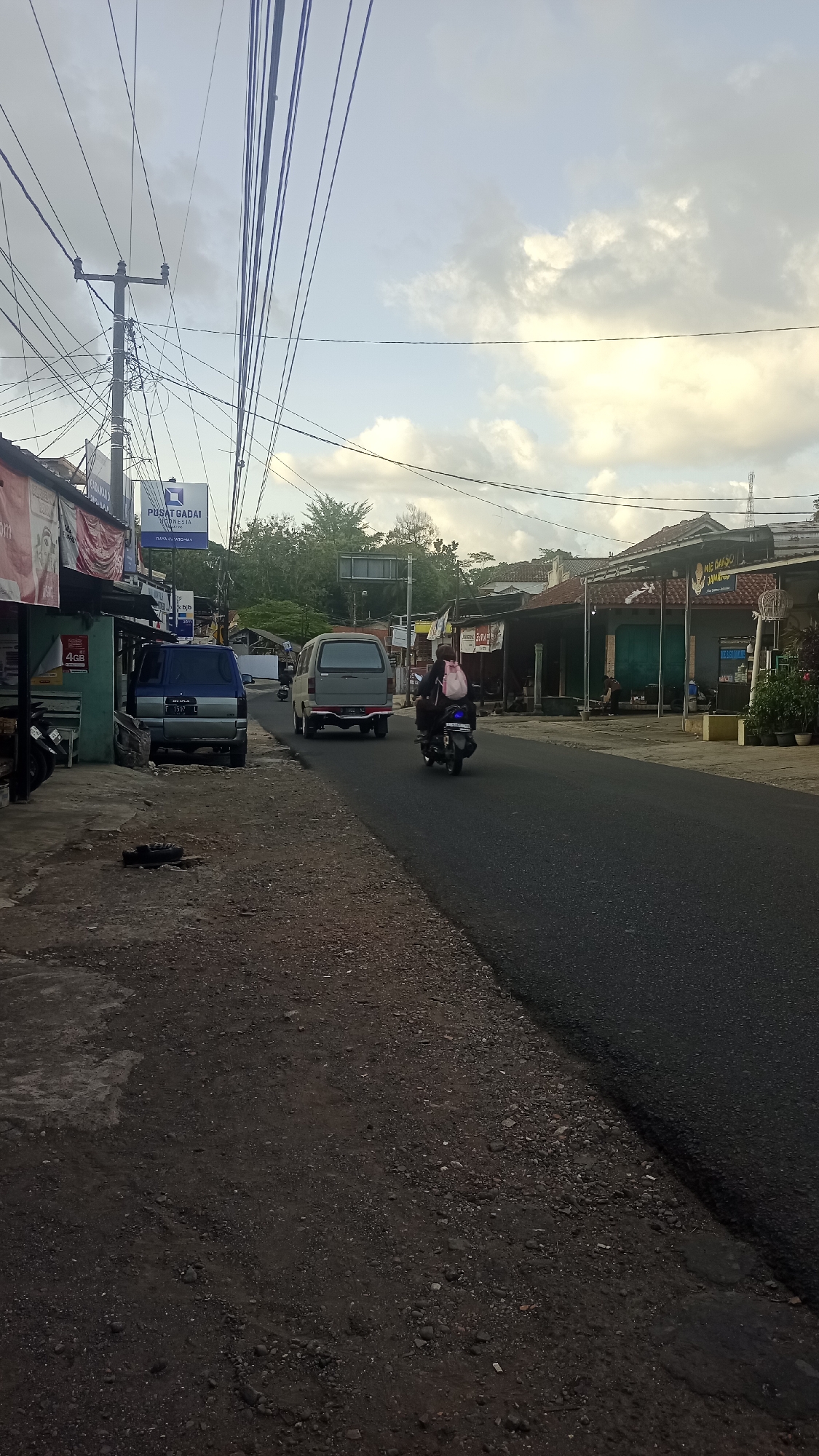 The image depicts a street scene in Pakemitan, Indonesia, under a partly cloudy sky. A paved road runs through the scene, with a gravel shoulder on the left. Several utility poles with a dense network of wires are visible. On the left side of the road, there is a row of small buildings and shops, including one with a blue sign that reads "PUSAT GADAI". A blue and silver utility vehicle is parked on the side of the road. Further down the road, a gray utility vehicle is driving away from the camera. A person riding a motorcycle, carrying a pink backpack, is also traveling away from the camera. On the right side of the road, more buildings with storefronts are present, including one with a sign that appears to say "MIE AYAM JAMUR". Trees and foliage are visible in the background. The overall atmosphere suggests a somewhat rural or suburban commercial area.