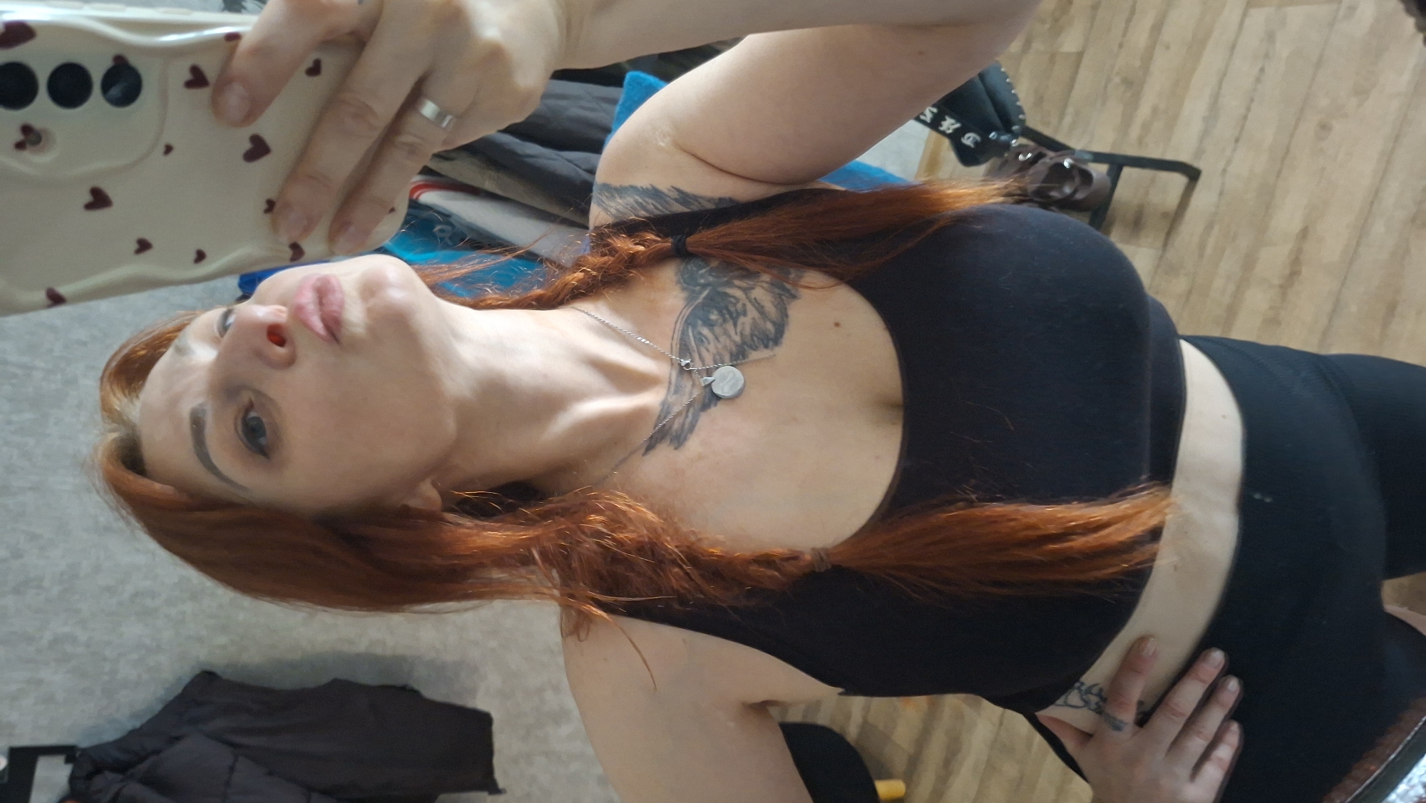 A woman with reddish-brown hair styled in two braids and wearing a black crop top and black shorts is taking a selfie in a room. She is looking up towards the camera, with her lips pursed. She has a tattoo on her chest and another on her left arm. A silver necklace with a circular pendant is visible. The background includes a wall with clothing hanging on it, a wooden floor, and furniture. This image was taken in a private residence in Myadzyel, Belarus.