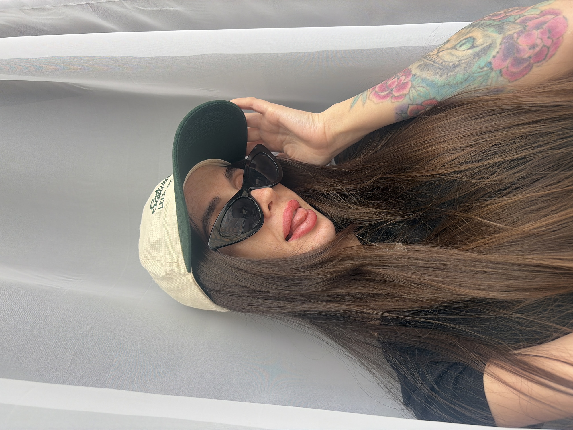 An indoor, chest-up image depicts a female individual in Saint Petersburg, Russia. She has long, straight, dark brown hair and is wearing a beige baseball cap with a dark green brim, featuring "Saturday Leisure" text in green script. She wears black cat-eye sunglasses and a black top, with a silver necklace visible. Her left arm displays a full sleeve tattoo of colorful floral patterns, including pink peonies, and a blue-teal animal face design. The individual is looking towards the camera, sticking her tongue out, which is covered in red lipstick, and is holding the brim of her cap with her left hand. The background consists of a plain, light-colored, vertically draped curtain or sheet.