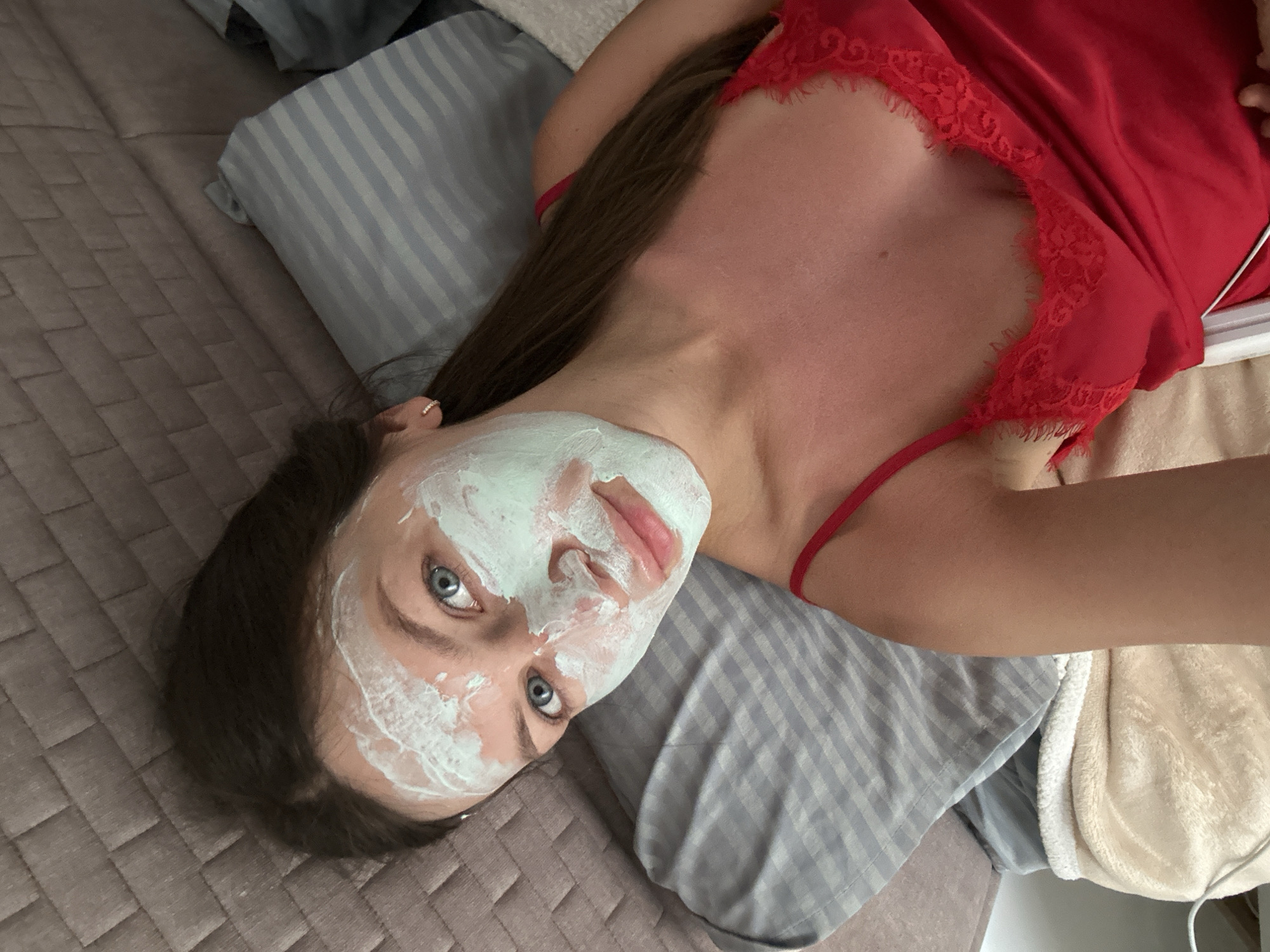 A woman is lying on a bed with a light blue and grey striped pillow under her head. She has a white and pale green facial mask on. She has blue eyes, brown hair, and fair skin. She is wearing a red satin top with lace trim. The bed has a quilted brown cover. The location cannot be determined from the image, but it is presumed to be Donji Pobori, Montenegro, based on the user's prompt.