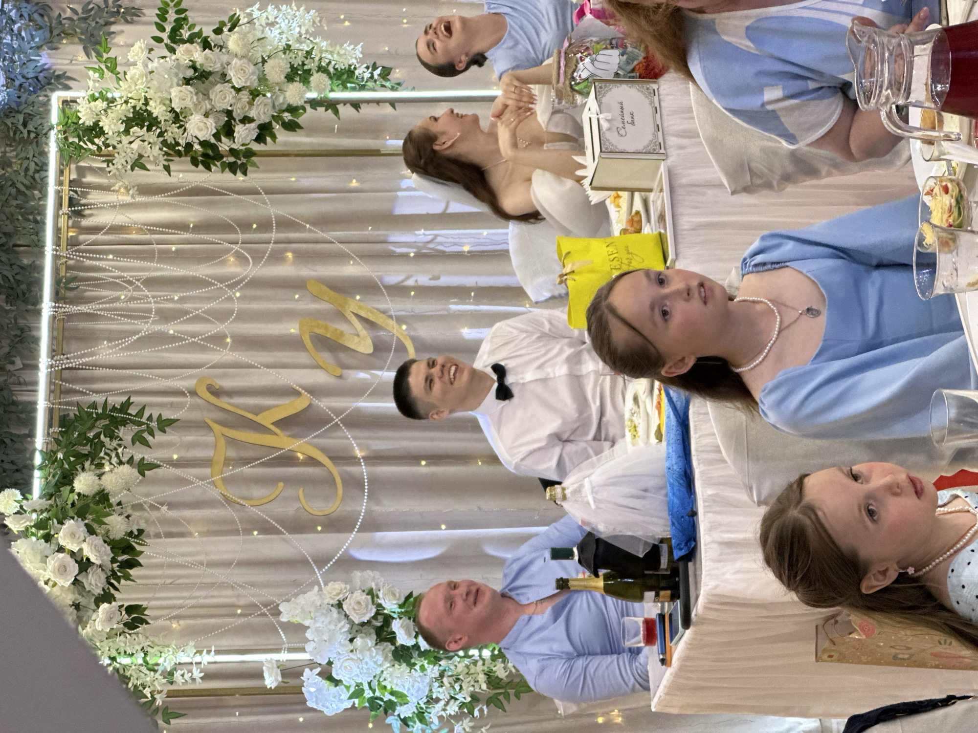 The scene depicts an indoor event, likely a wedding reception, at a venue in Chaikovsky, Russia. The background features a light beige pleated curtain serving as a backdrop, adorned with two golden rectangular frames or arches decorated with lush artificial white flowers and green foliage. Draped across the backdrop are numerous strands of white pearls, creating a web-like pattern, interspersed with small, warm white fairy lights. Centrally positioned on the backdrop are two golden Cyrillic letters, "К У". A long table, covered with a white pleated tablecloth, occupies the midground and foreground. Seated from left to right in the midground are four individuals. The leftmost is an adult male with light skin and short light brown hair, wearing a light blue Mandarin-collar shirt and a gold chain; he smiles towards the right. In front of him are a glass with a red beverage, a smartphone, a pen, two dark green bottles (one with a miniature white sheer dress draped over it), and a partially visible food plate. To his right is a young adult male with dark hair, wearing a white dress shirt and a black bow tie, smiling widely towards the right. Next is a young adult female, presumably the bride, with long dark brown hair and a white veil, wearing a white off-the-shoulder gown and a pearl necklace; she smiles towards the right with her hands clasped. In front of her are a yellow gift bag, a small white wooden box labeled "Семейный банк" (Family Bank), and a large clear jar filled with colorful items. The rightmost adult is a young female with dark hair pulled back, wearing a light blue top; she is laughing heartily, looking towards the right. In the foreground, two girls are seated. The leftmost girl has long dark brown hair, wears a light blue top, a pearl necklace, and pink flower earrings; she looks towards the left with a pensive expression. To her right is another girl with long dark brown hair, wearing a light blue long-sleeved dress and a pearl necklace with a small pendant; she looks towards the right. Partially visible on the far right foreground is an adult female wearing a light blue and white patterned top. On the table in the foreground are clear glasses, a small bowl of food, and a clear pitcher containing a red beverage. The overall atmosphere is celebratory, with most adults displaying smiles or