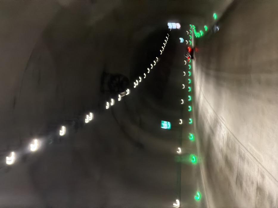 The image depicts the interior of a vehicular tunnel, identified as being located in Terjola, Georgia. The perspective is from within a moving vehicle, looking forward. The tunnel features light grey, possibly concrete, walls and ceiling, forming an arch. A series of bright white overhead lights illuminate the tunnel's upper section, appearing as blurred, elongated 'U'-shapes due to motion. Along the base of both tunnel walls, a continuous line of illuminated green markers delineates the roadway. The road surface itself is paved, grey, and appears slightly reflective or damp. In the mid-distance, a dark-colored passenger vehicle is observed, its red taillights distinctly visible, indicating its position ahead of the vantage point. Two blurred, green-lit rectangular signs are present on the left tunnel wall, their specific content indecipherable. No people are visible. The scene suggests active transit through the tunnel.