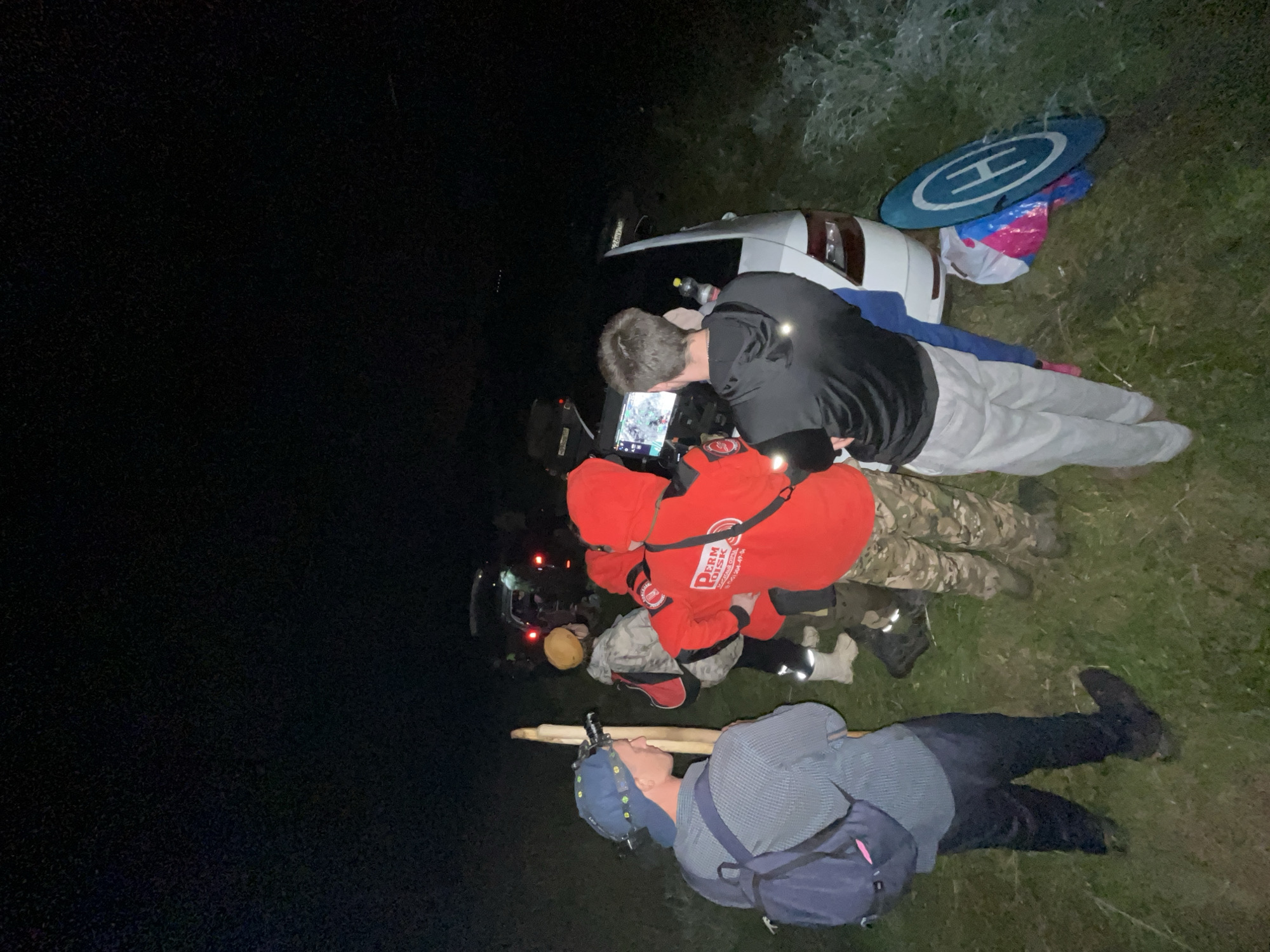 A group of at least six individuals are gathered in a grassy, outdoor area at night, near several vehicles, one of which is a white car with its trunk open. The scene appears to be in or near Badashka, Russia, given the context. Some individuals are wearing headlamps or other lighting devices. One person in the foreground wears a blue baseball cap and a dark backpack, holding a long wooden pole. Another person is dressed in a bright red hooded jacket with "PCRN RUSSIA" printed on the back. Several individuals appear to be looking at a screen, possibly a tablet or monitor, being held by one person. A blue and white helipad lies on the ground next to the white car, along with some brightly colored bags. The surrounding environment is dark, with only the lights from the vehicles and headlamps illuminating the immediate area, suggesting a search or rescue operation is underway. The ground is covered in grass and possibly some weeds.