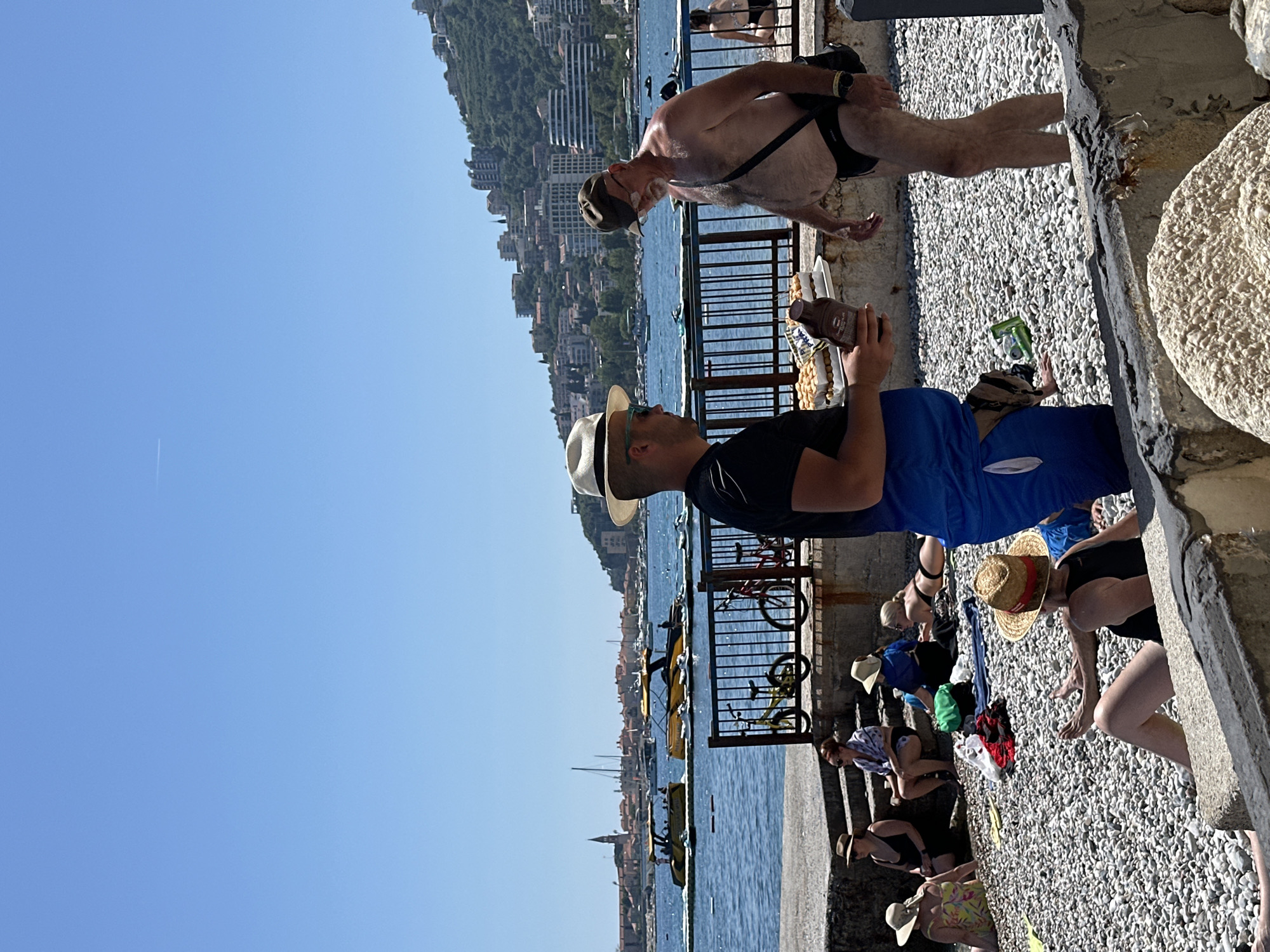 In Budva, Montenegro, on a sunny day at the beach, several individuals are present. In the foreground, two men are interacting. One man, wearing a white fedora, sunglasses, and a black t-shirt with blue shorts, holds a bottle of chocolate syrup and stands next to a tray of pastries. The other man, wearing a brown cap, glasses, and black swim trunks, stands shirtless with a dark strap across his chest. Several people are seated and lying on the pebble beach behind them, some wearing hats and swimwear. In the background, the blue Adriatic Sea is visible, dotted with yellow jet skis and boats. Beyond the water, the city of Budva is spread out, with buildings nestled against a lush green hillside. A black metal fence runs along the edge of the beach promenade.