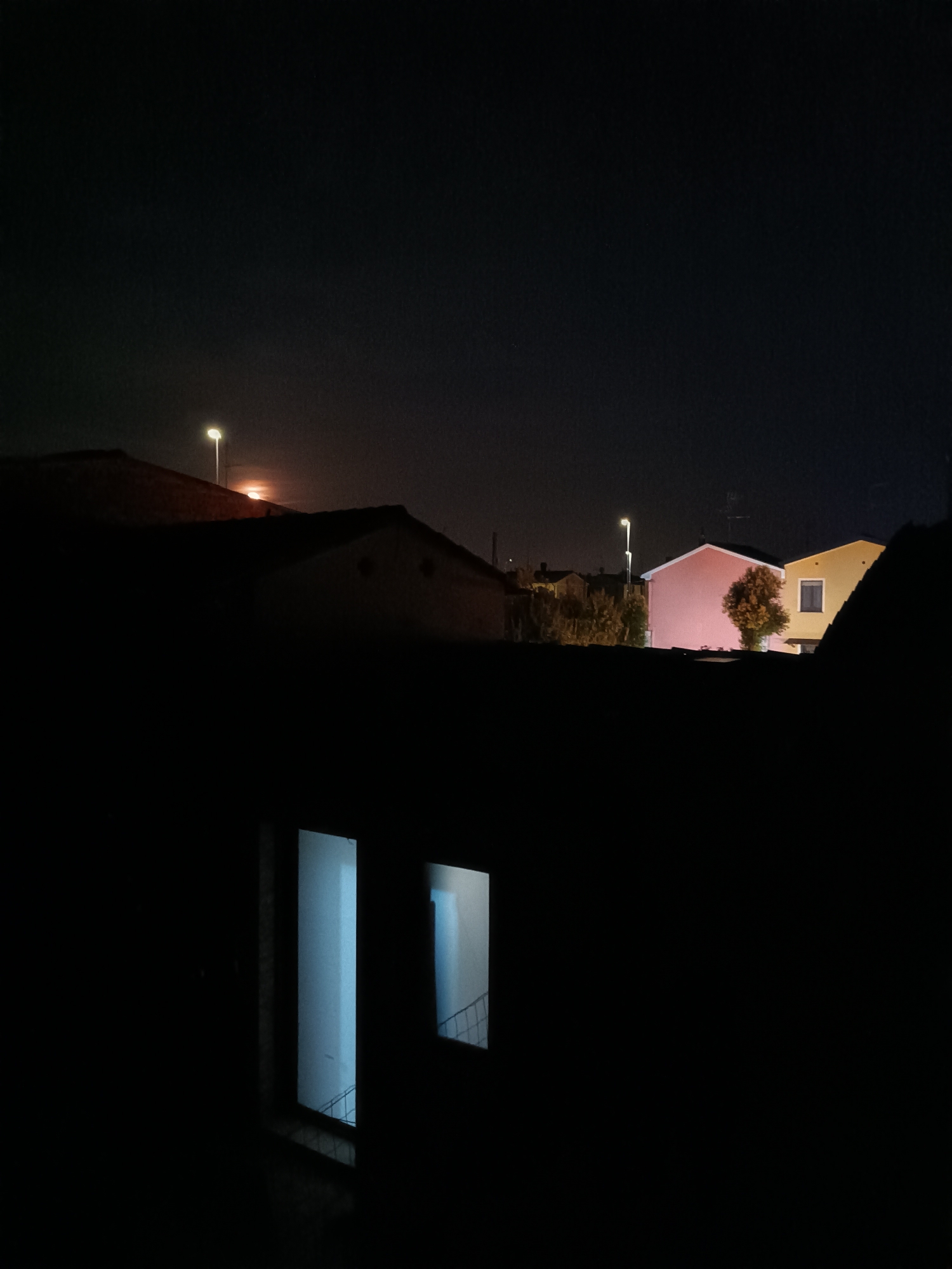The image presents a tranquil nighttime scene captured from an elevated perspective, overlooking a residential area in Viadana, Italy. The foreground is dominated by the dark silhouettes of rooftops and buildings, with the night sky appearing largely black.
In the mid-ground, a cluster of houses emerges from the darkness. Notable among them are a house with a pale pink facade and another with a yellow/ochre exterior, featuring a single dark window. A small tree with light-colored foliage stands directly in front of the pink house. The scene is illuminated by several sources: two bright, white-yellow streetlights pierce the darkness, one appearing behind a foreground rooftop, and another further back, casting light upon the pink house.
A prominent reddish-orange glow, indicative of a distant moon or significant atmospheric phenomenon, is visible in the sky above one of the foreground rooftops, near the higher streetlight.
In the lower foreground, two windows from a closer building reveal interiors lit with a cool, ethereal bluish light. Through the left window, a structure resembling a railing or staircase is faintly discernible. The image captures a static moment; no specific activity or event is taking place. The overall impression is one of quiet stillness, contrasting the deep shadows with the various sources of light that punctuate the dark canvas of the night. There is no visible text within the image.