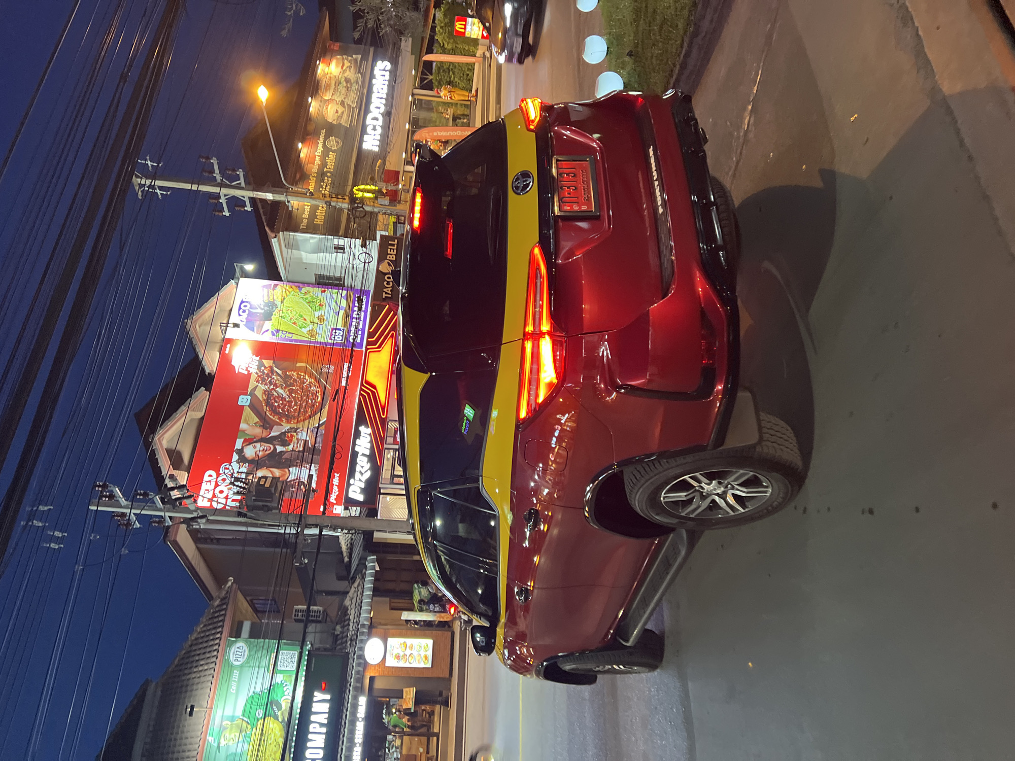 The image depicts a red and yellow taxi in Baan Bo Phut, Thailand, parked on the side of a street at dusk. The taxi, identified as a Fortuner, has its rear facing the camera, revealing a license plate with Thai characters and numbers. In the background, various businesses are illuminated, including Pizza Hut, Taco Bell, and McDonald's, suggesting a commercial area. Power lines crisscross the sky above the buildings. The street appears to be paved, and a grassy area with decorative globe lights is visible to the right of the taxi. The overall scene is characteristic of a bustling urban environment at night, with commercial signage and transportation infrastructure dominating the view.