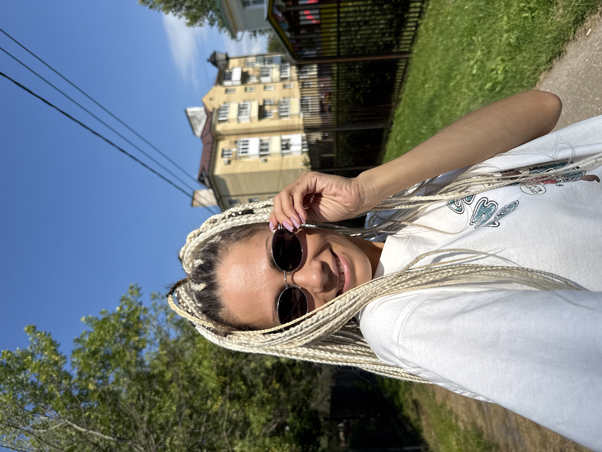 A woman with blonde braids is taking a selfie outdoors on a sunny day. She is wearing a white t-shirt and sunglasses. Behind her, a yellow apartment building is visible, along with trees and a fence. The location is in Rusanovo, Russia. The image has a shallow depth of field, with the woman in sharp focus and the background slightly blurred.