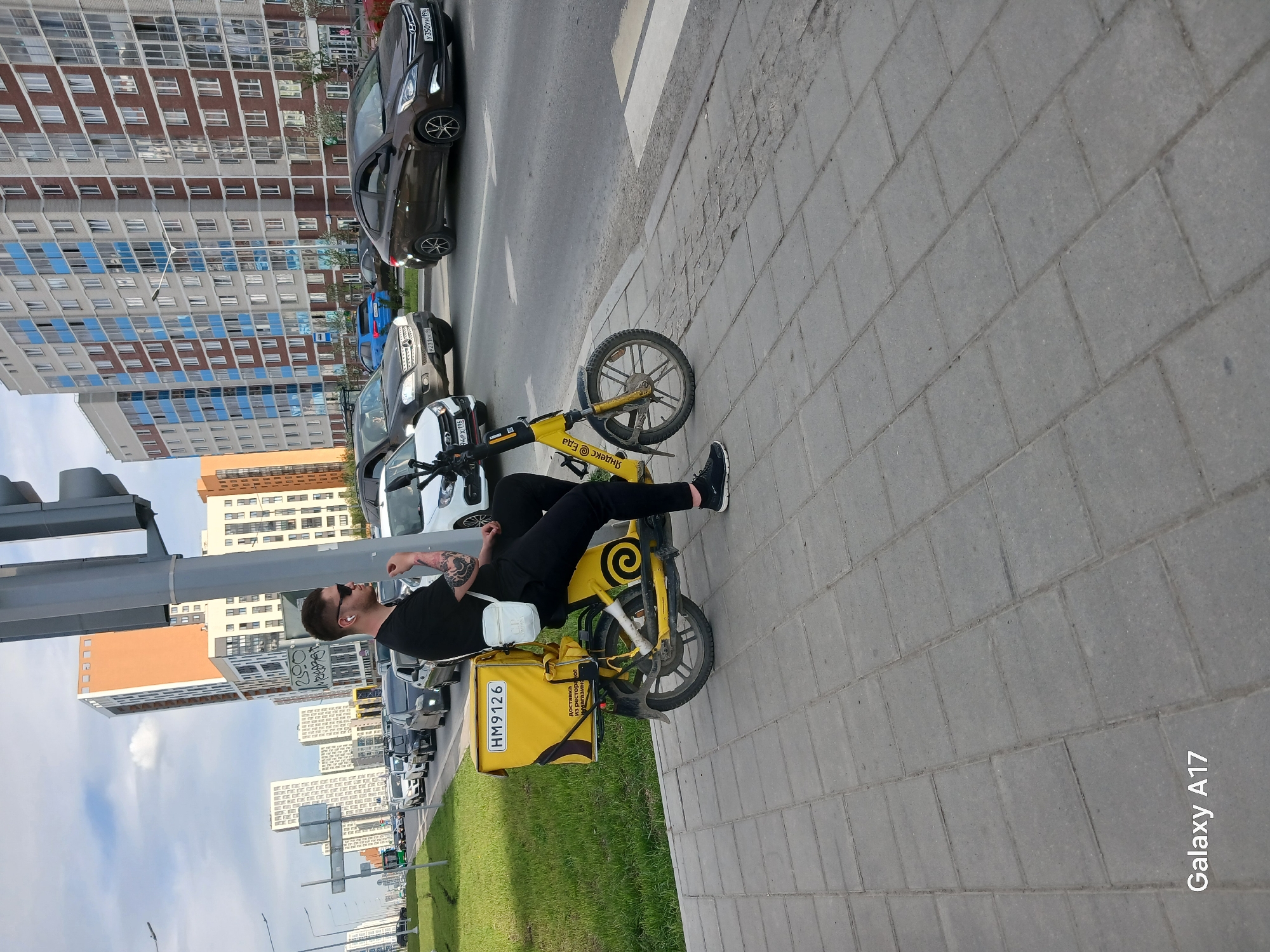 A man is sitting on a yellow electric bicycle with a delivery bag on a paved sidewalk. He is wearing black clothes, sunglasses, and appears to have a tattoo on his arm. The delivery bag has the license plate "HM9126" and the words "Яндекс Еда", which translates to "Yandex Food", indicating that the man is a delivery driver for the Yandex food delivery service.
In the background, there is a city street with several cars lined up. There are tall apartment buildings on either side of the street. A traffic light is visible on the left side of the image. There is grass in between the sidewalk and the street with several trees.
The text "Galaxy A17" is printed on the bottom right corner of the image, suggesting that the photo was taken with a Samsung Galaxy A17 phone. Based on the "Яндекс Еда" branding and license plates, the location is most likely Yekaterinburg, Russia.