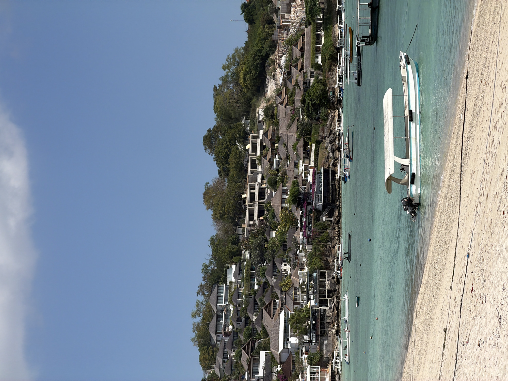 A long shot captures a bay in Nusa Penida, Indonesia, on a sunny day. The water is a clear, turquoise color, with several boats anchored in the bay. One boat, a white and blue motorboat, is closer to the shore, where a sandy beach stretches across the foreground. Behind the bay, a hillside is covered with a collection of buildings, many with traditional thatched roofs, nestled among lush green trees. The sky above is a clear, bright blue, with a few wispy clouds near the top edge of the frame.