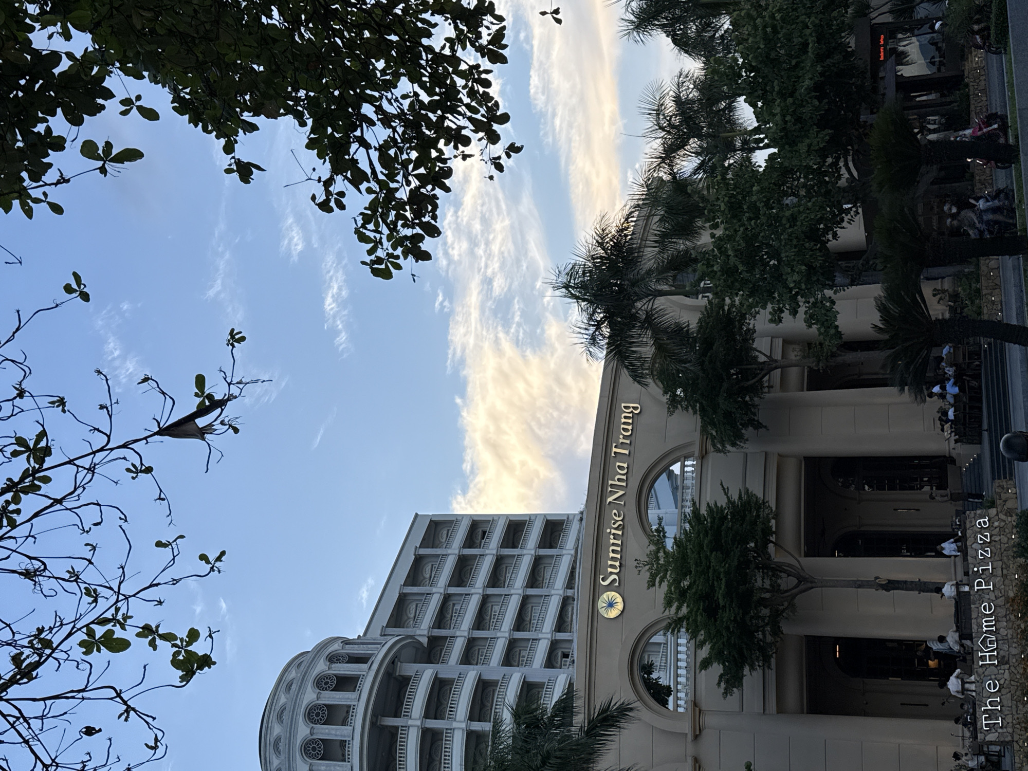 In Nha Trang, Vietnam, the Sunrise Nha Trang hotel is visible with its grand architecture, featuring a domed section and multiple balconies. In front of the hotel, the restaurant "The Home Pizza" is displayed on a stone wall. Palm trees and other foliage surround the building. The sky above is blue with scattered clouds. Several people are seated at outdoor tables in front of the restaurant, and a few individuals are on motorcycles on the street.