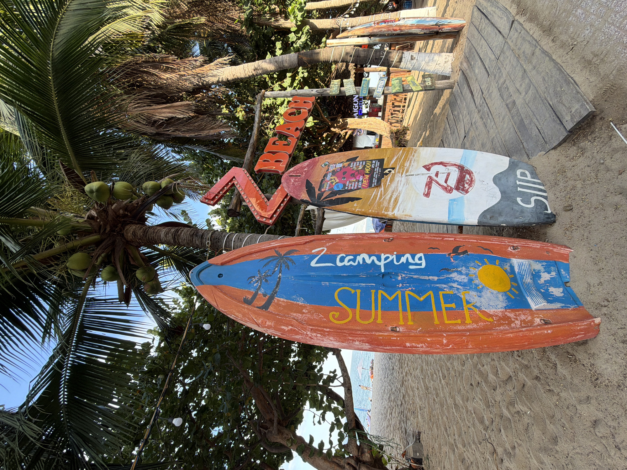The image shows a beach scene in Nha Trang, Vietnam, featuring several surfboards and palm trees. In the foreground, two colorful surfboards are propped up on the sandy beach. The surfboard on the left is primarily orange with blue and yellow accents, and it has the word "SUMMER" written vertically in yellow. The surfboard on the right is white with red and black graphics, and it has the word "SIIP" written horizontally at the bottom.
Behind the surfboards, a wooden pathway leads towards a beach bar or restaurant area, indicated by a wooden structure with tables and chairs. A prominent red neon sign spelling out "BEACH" in large letters is visible above the pathway. Several smaller directional signs are mounted on poles, including one for "SAIGON" and another for "DETOX."
Tall palm trees with coconuts are scattered throughout the scene, their fronds casting shadows on the beach and the wooden structures. In the background, the ocean is visible, with a few people and beach umbrellas on the sand. The sky is clear and blue, suggesting a sunny day. The overall atmosphere is that of a tropical beach resort.