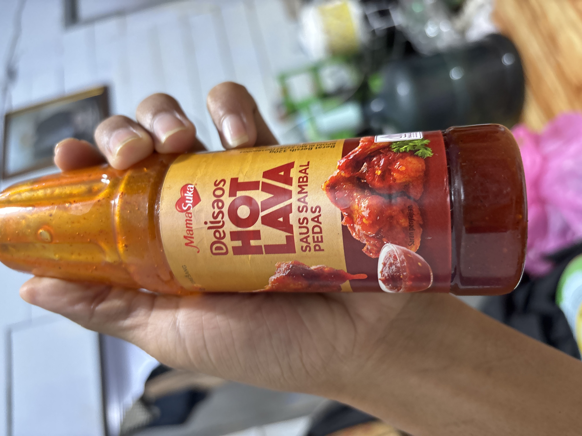 A hand holds a plastic bottle of "MamaSuka Delisaos Hot Lava Saus Sambal Pedas" hot sauce. The bottle is transparent, revealing the reddish-orange sauce. The label is a light yellow color, with red and dark brown accents. The hand appears to be lightly tanned. The background is blurry, but suggests an indoor environment. The location is Mideuenjok, Indonesia.