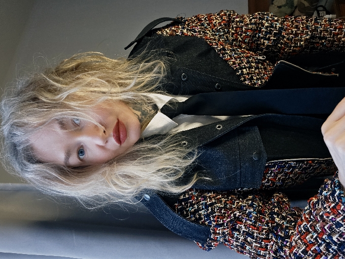 A blonde woman with light eyes and red lipstick is seen wearing a white shirt with a black tie and a tweed and denim jacket. She has a tattoo on her neck. The jacket has a rough, multi-colored tweed section contrasted with a dark blue denim fabric. She is lying down on a grey surface. Her pose is somewhat tilted, adding a unique angle to the shot.