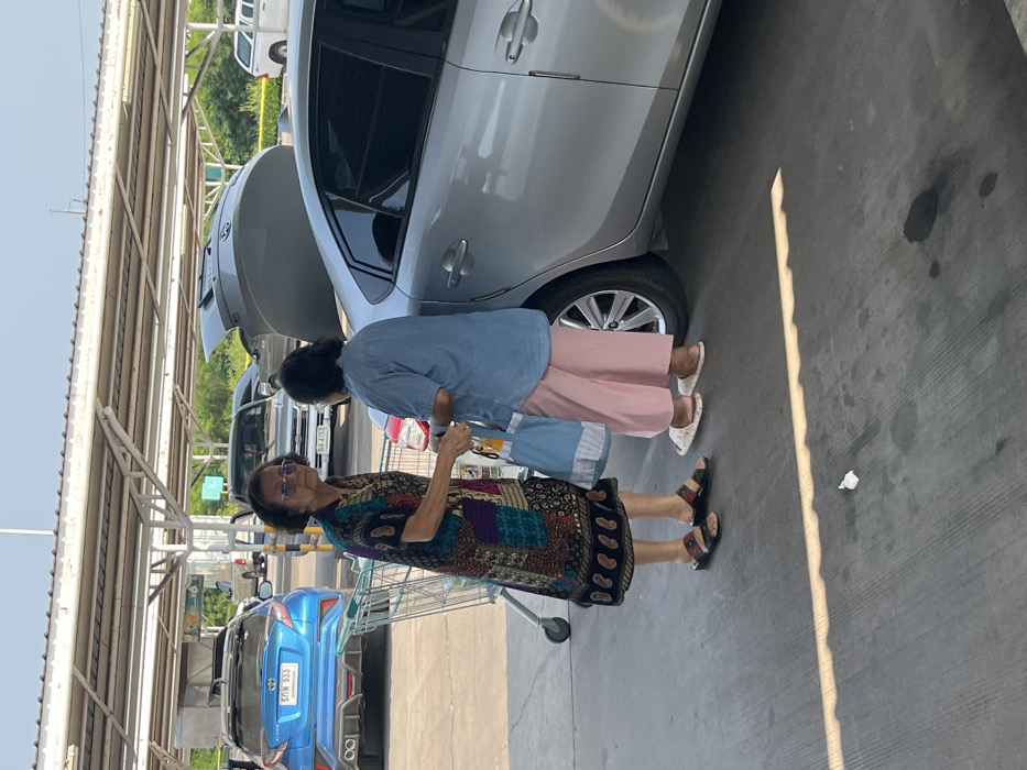 The scene depicts two elderly women engaged in an exchange within a covered outdoor parking area, likely associated with a commercial establishment in Bang Pu Subdistrict Municipality, Thailand.
The woman on the left, wearing dark sunglasses, a multi-patterned knee-length dress, and dark sandals, stands beside a metal shopping cart. Her right arm is extended towards the second woman. The woman on the right, observed from her back-right side, wears a light blue short-sleeved top, light pink wide-leg trousers, and white slippers. She holds a light blue reusable bag with white stripes and faces the first woman.
The interaction takes place adjacent to a silver four-door sedan, which is parked with its trunk open, indicating items are being loaded or unloaded. In the background, a blue Toyota C-HR with Thai license plate "5กค 933" is parked to the left, and a silver pickup truck with license plate "3ข 4153" is partially visible further back. The ground is concrete, showing some stains and a discarded piece of white paper. Overhead, a light-colored, open-frame canopy provides partial shade, casting distinct shadows on the ground under bright daylight conditions. Green foliage is visible in the distant background under a clear sky.