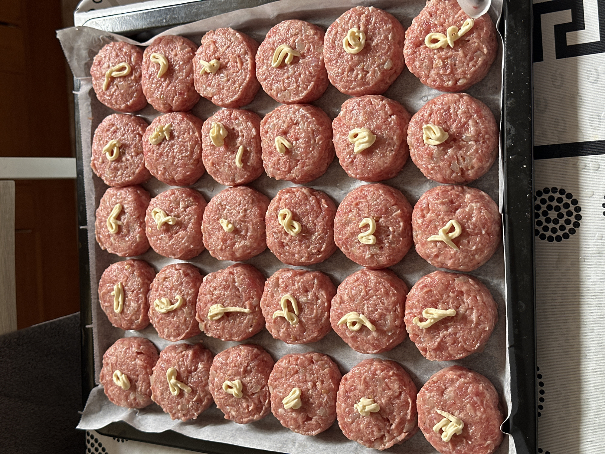 A close-up, overhead view shows a baking tray lined with white parchment paper, filled with approximately 30 raw, flattened hamburger patties. Each patty has a small dollop of white mayonnaise on top, some arranged in swirls or lines. The patties are arranged in neat rows and columns. The background is blurred, suggesting an indoor setting with wooden furniture. The overall scene appears to be a food preparation area.