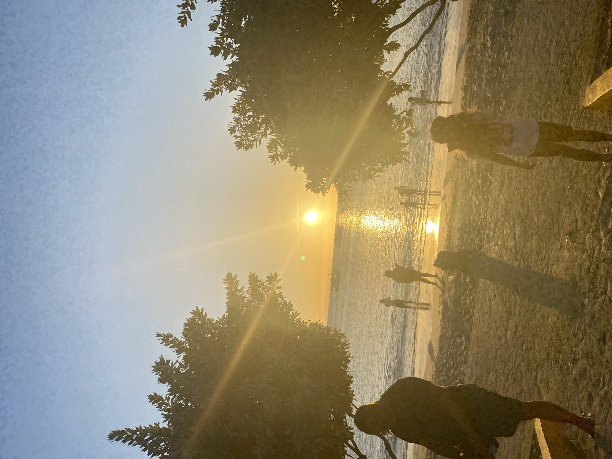 The image shows a beach scene in Juan Griego, Venezuela at sunset. The sun is low on the horizon, casting a golden glow on the water and sand. Silhouettes of people are visible along the shoreline and in the water. Trees frame the view from the sides, and individuals are standing on a ledge observing the sunset.
