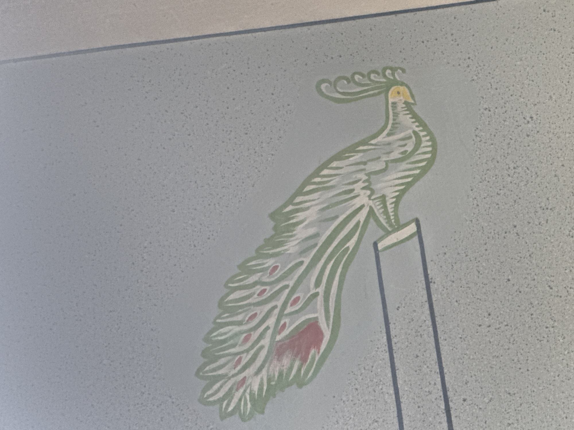 The image shows a mural of a stylized peacock painted on a textured, light blue wall in Kosh, Armenia. The peacock is depicted in profile, facing left, with a crested head, a long, fanned tail, and a single vertical post it is perched upon. The artwork uses green and white for the bird's body and feathers, with accents of pink and yellow for details such as the eyespots on its tail and its beak. The wall itself has a speckled appearance, and a shadow from an overhead light source casts a faint line across the top of the image. The overall impression is that of a decorative element within an interior space.