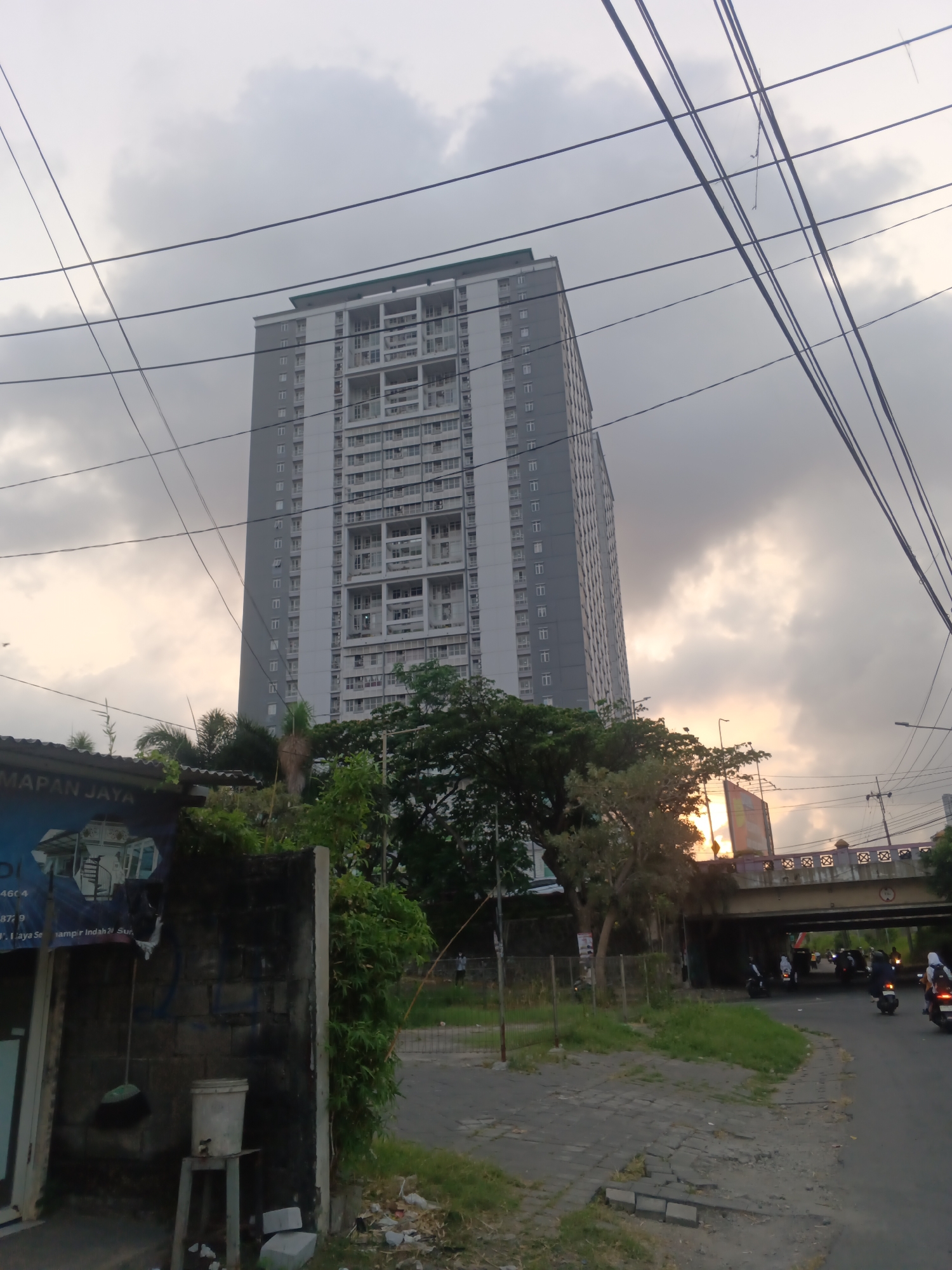 The image depicts an outdoor scene in Surabaya, Indonesia. Dominating the frame is a tall, modern apartment building with a light gray facade and numerous windows. Power lines crisscross the sky in the foreground. To the left of the building, a portion of a building with a sign that reads "MAPAN JAYA" is visible, featuring blue and white graphics and what appears to be an elevator. In front of this building, a concrete wall with some graffiti can be seen, along with a broom leaning against it and a bucket on a stand. Dense green foliage and trees partially obscure the base of the apartment building and extend towards the right side of the image. A road runs through the scene, with several motorcycles and riders visible. An overpass stretches across the road in the middle ground, with traffic passing underneath. The sky is overcast with patches of sunlight breaking through, suggesting it may be late afternoon or early morning. The overall environment appears to be an urban setting with a mix of residential, commercial, and infrastructural elements.