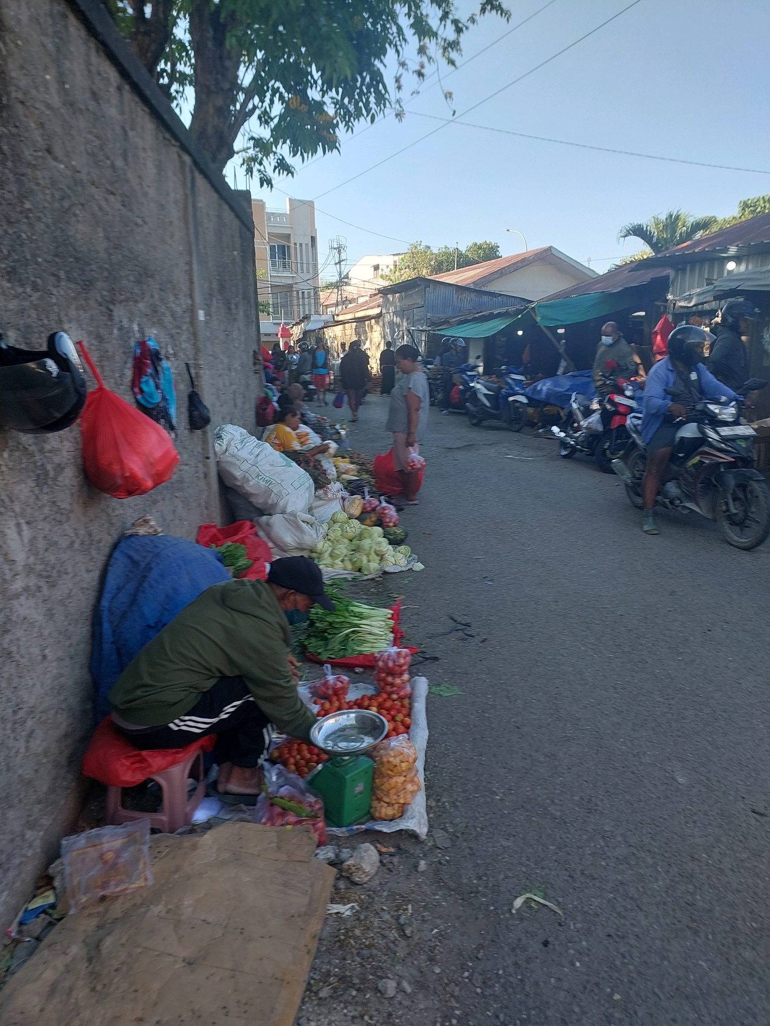 The image depicts a street market scene in Kupang, Indonesia, likely in an open-air setting with modest structures. A vendor, wearing a green jacket and black pants with white stripes, is crouched down next to a display of fresh produce. They are seated on a red stool. The produce includes tomatoes, leafy greens like spinach or kale, and cabbage. A metal bowl and a green scale are also visible among the goods. In the foreground, a brown tarp is spread out on the ground, and to the left, a rough concrete wall serves as a backdrop. Bags, including a large red one, and other items are hanging from the wall. Behind the vendor, a large blue tarp is draped over something. The market appears to be active, with several motorcycles parked along the street and people browsing or walking by. The overall environment suggests a typical urban market setting in a tropical climate, with clear daylight and a blue sky.