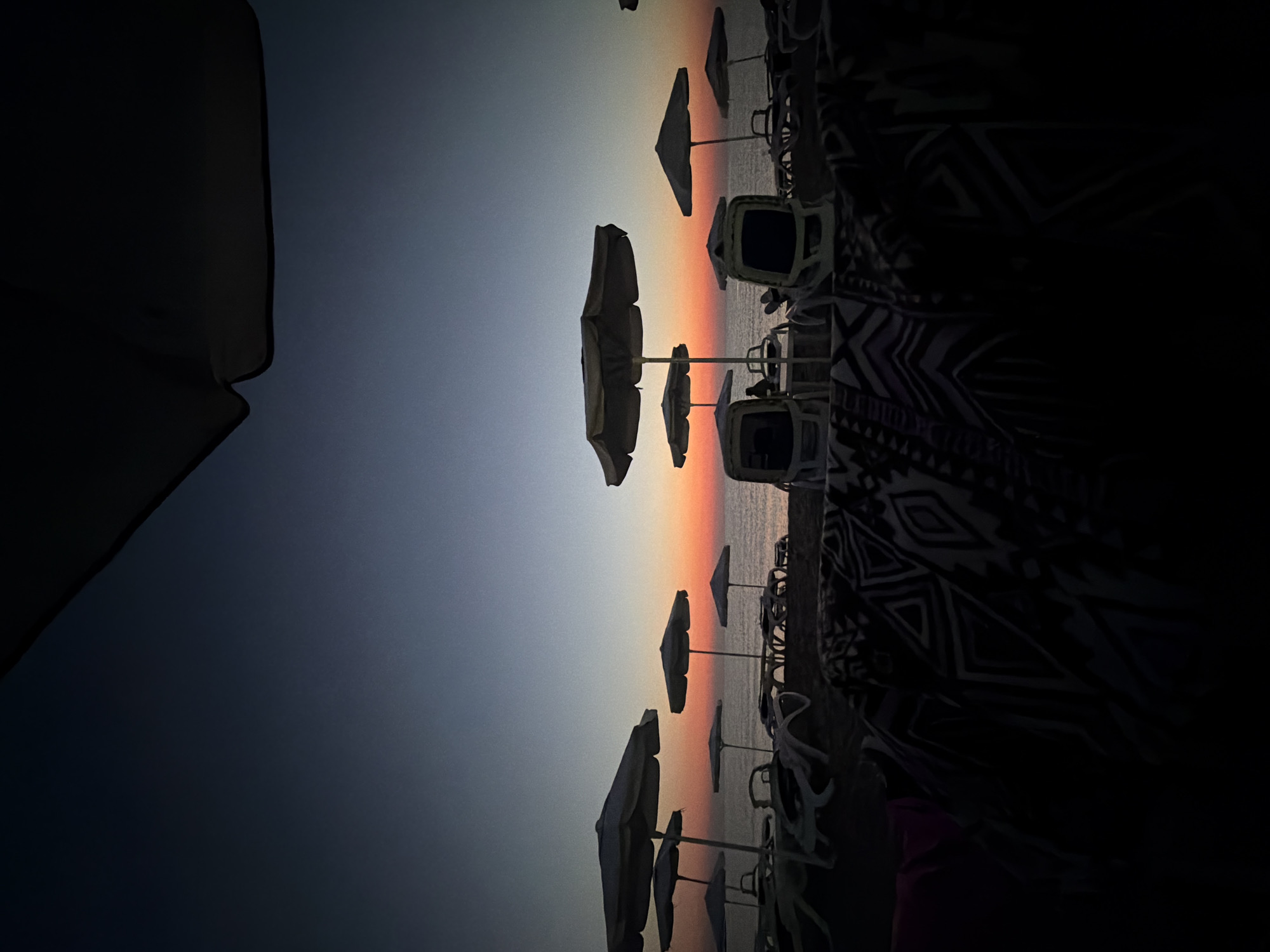 The image depicts a twilight scene on a beach in Lozenets, Bulgaria. The sky displays a gradient of colors, transitioning from deep blue at the top to a vibrant orange and pink hue near the horizon, indicating either sunrise or sunset. Numerous empty lounge chairs and beach umbrellas are arranged on the sand, suggesting a public beach that has emptied for the day or is preparing for its busiest hours. The sea is visible in the background, calm and reflecting the sky's colors. In the foreground, dark patterned blankets or towels are draped over what appear to be more lounge chairs, obscuring some of the lower portion of the scene. The overall impression is one of peacefulness and tranquility at the end of a day.