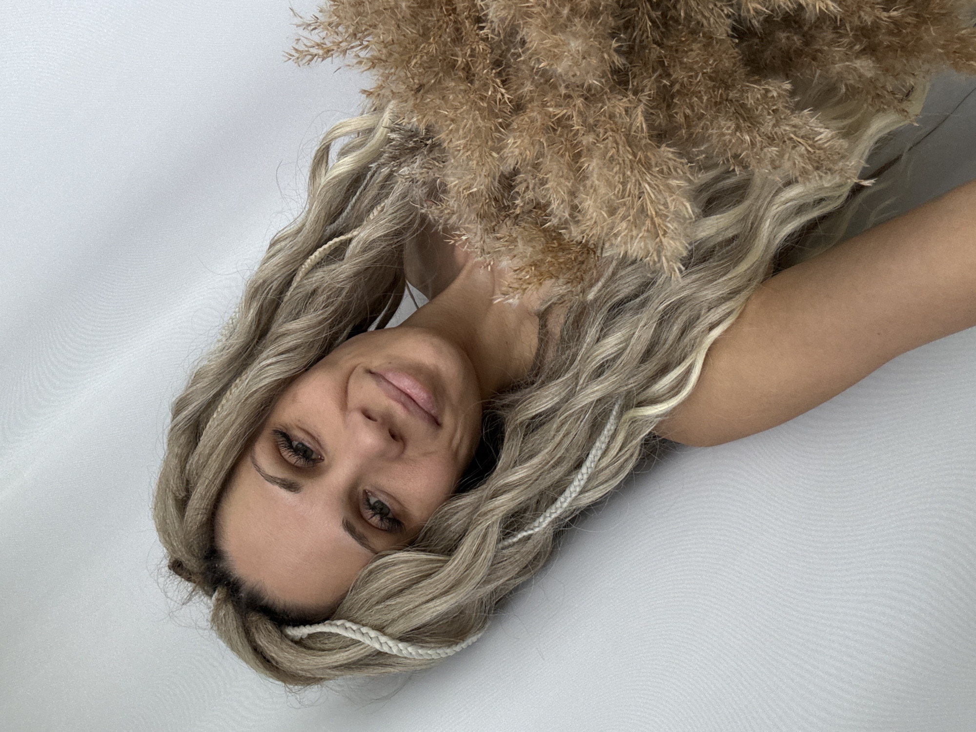 A woman with long, wavy, blond hair, adorned with white braided accents, is holding a bouquet of dried pampas grass. Her face is angled towards the camera, and her body is partially visible on a light grey or white surface, possibly fabric or a wall. The location in the city of Rusanovo, Russia, is not evident in the image.