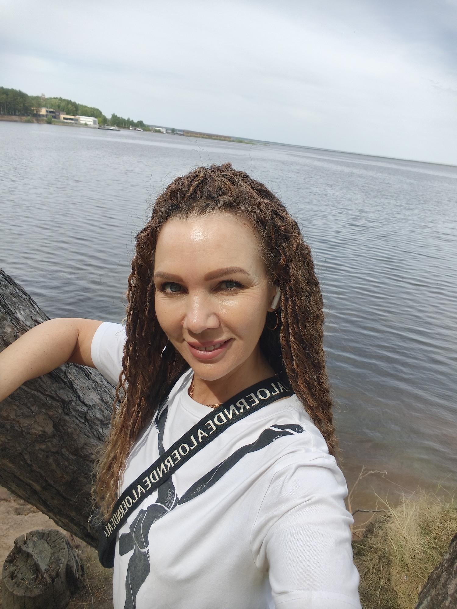 The image shows a woman with long, curly brown hair standing outside near a large body of water. She is leaning against a tree trunk, and the water is visible in the background. The location is described as Unknown, Unknown.