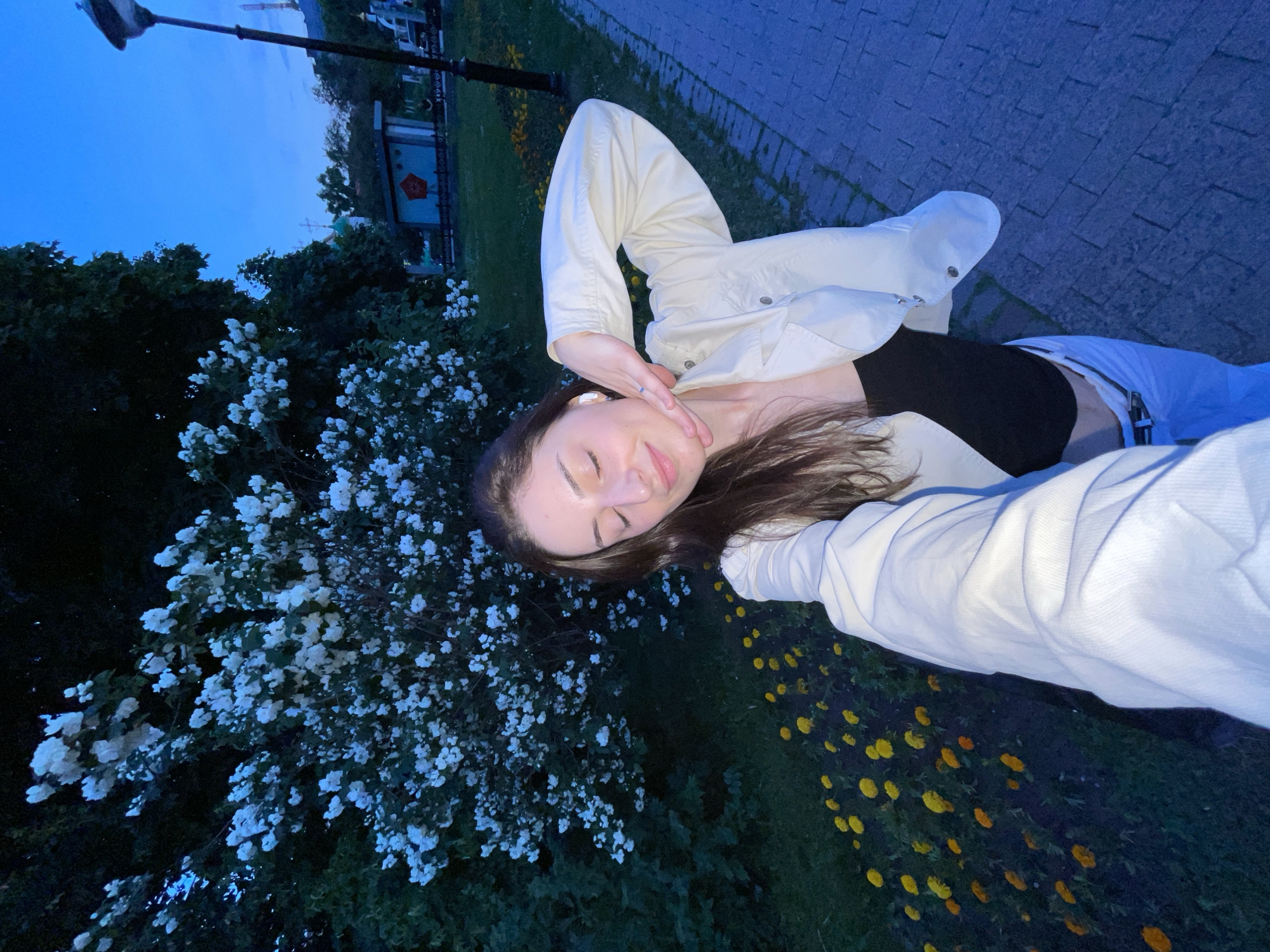 A young woman with long dark hair and closed eyes takes a selfie outdoors in what appears to be a park at dusk. She is wearing a white jacket over a black top and white pants. Her left hand is raised, touching her chin. Behind her, a large bush with abundant white flowers is visible, with a line of yellow flowers along the base. To the right, a paved walkway leads away from the viewer, and a lamppost stands beside it. In the background, trees, a building with a red star emblem, and a distant structure with a dome are discernible. The sky is a deep blue, indicating evening. The scene is likely located in Moscow, Russia, given the architectural style of the building with the red star.
