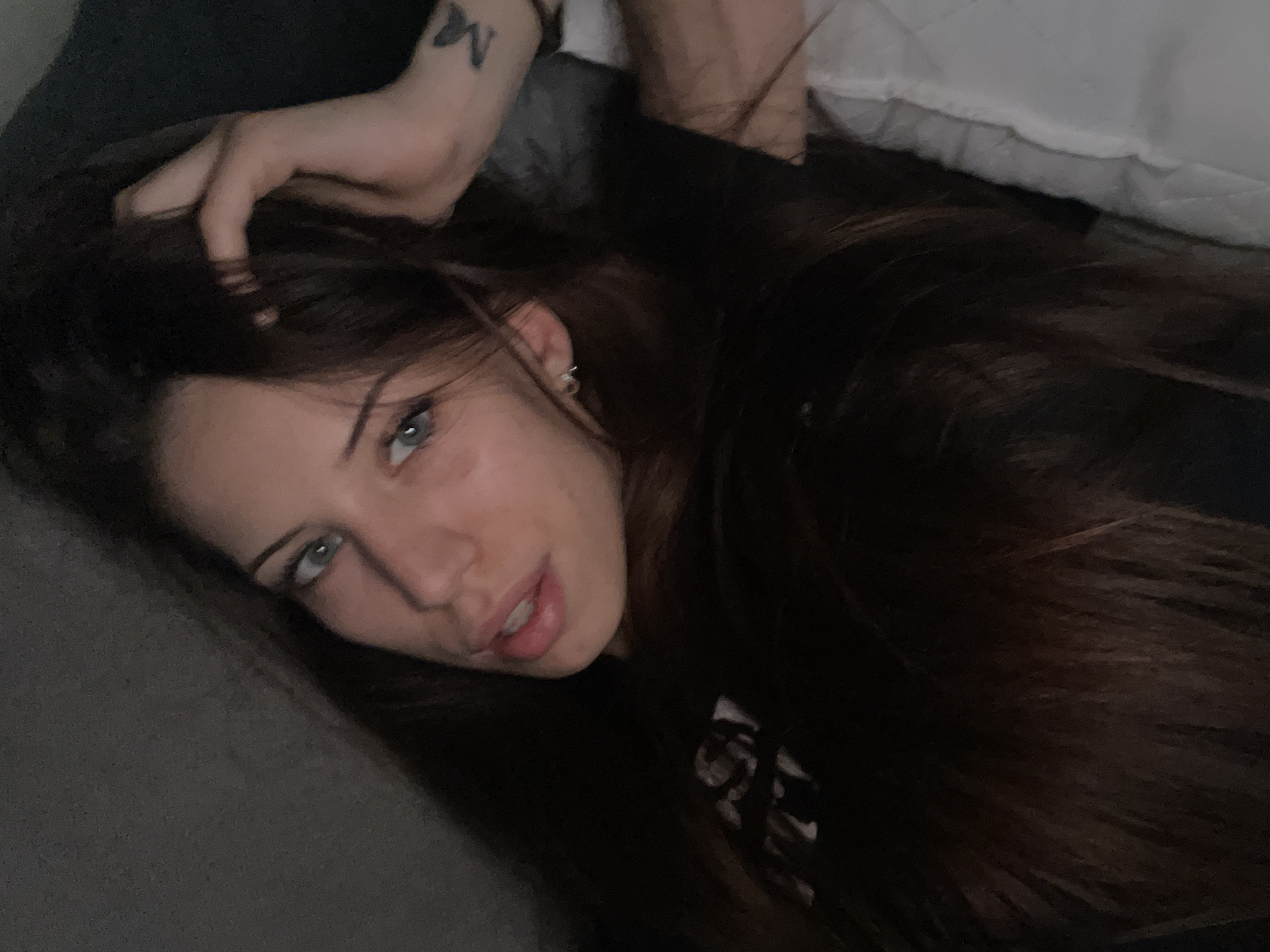 A young woman with long, dark brown hair and blue eyes is lying down, looking at the camera. She is wearing a black t-shirt. Her left hand is resting on her head, and she has a small tattoo of a butterfly on her left forearm. The background appears to be a dark, textured surface, possibly a wall or headboard. The lighting is dim, creating a soft, intimate atmosphere. There is a white, quilted surface to the right of the woman, possibly a pillow or blanket.
