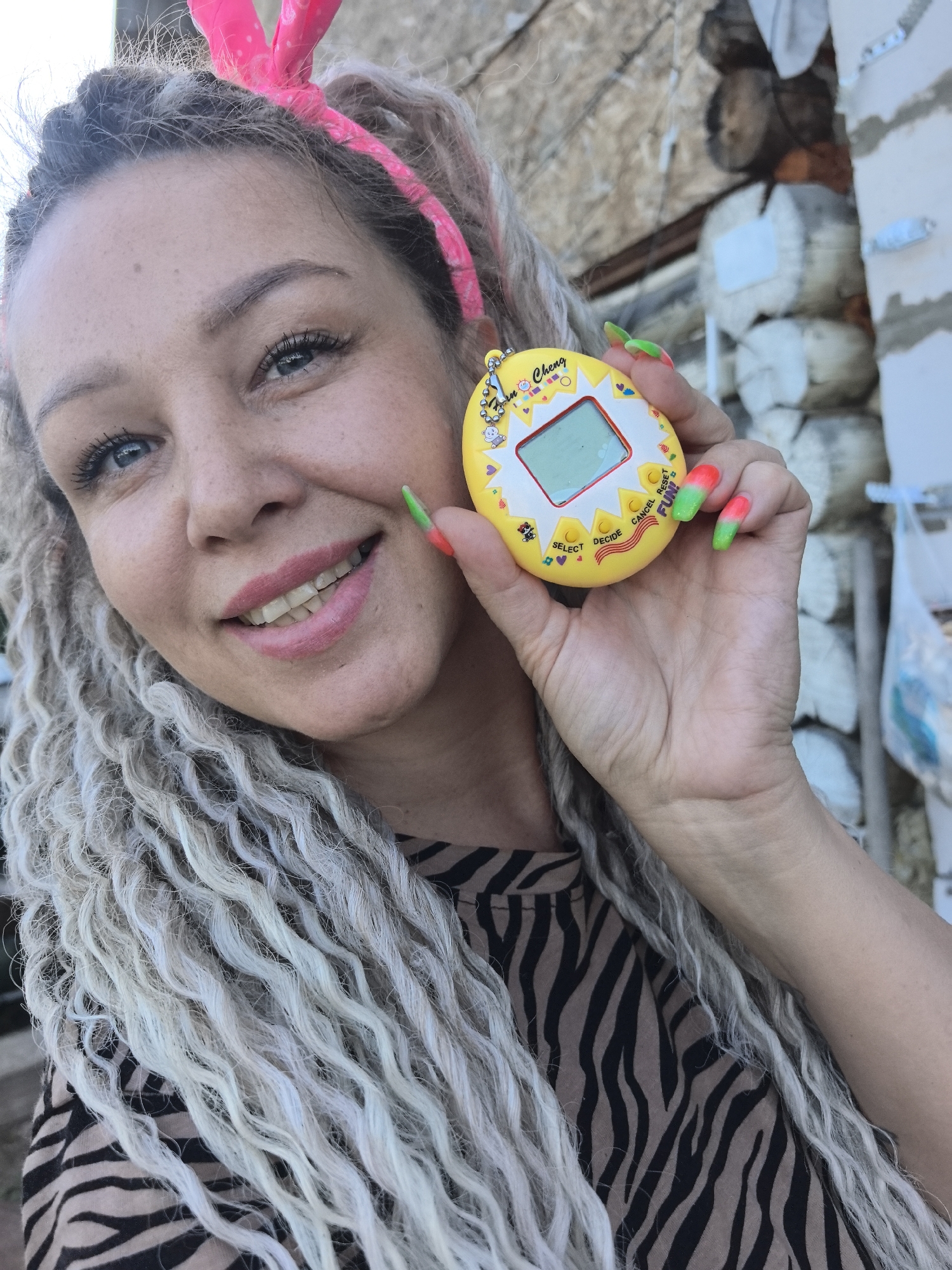 The image is a close-up selfie of a woman holding a yellow and white electronic toy, likely a Tamagotchi. She is smiling and looking at the camera. She has light skin, blue eyes, and light gray braided hair with a pink headband. She is wearing a black and brown zebra-striped shirt. Her fingernails are painted with a red-to-green ombre design. The background is blurred, but it appears to be an outdoor setting with a log structure partially visible. The location is Noviy, Russia.