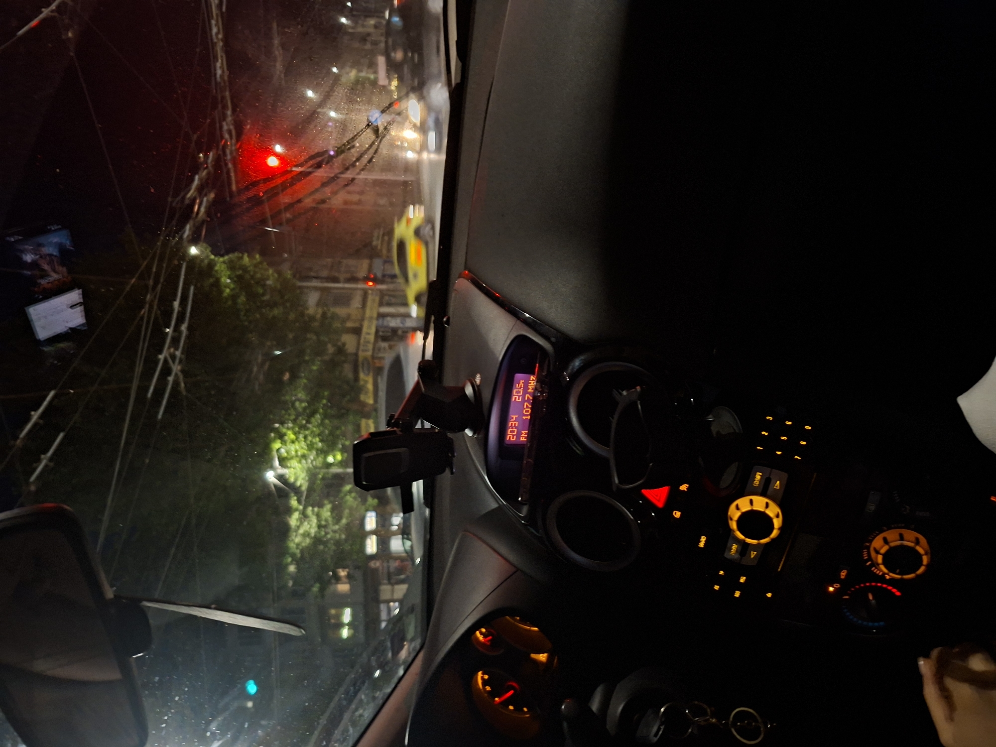 Here's a forensic description of the image:
**Setting:** Interior of a vehicle, likely a car, at night in the city of Pleven, Bulgaria. This is inferred from the display showing "107.7 MHz," a frequency used by local FM radio stations in Pleven. The view is through the windshield.
**Environment:** Rain or moisture is visible on the windshield. City street with buildings and trees, illuminated by streetlights and traffic signals.
**Objects:**
* Vehicle dashboard with illuminated buttons, gauges, and displays.
* Mobile phone holder mounted on the dashboard, with a mobile phone inside.
* Rearview mirror.
* City street scene visible through the windshield with buildings, trees, streetlights, and a yellow car/taxi.
* A billboard is on the left.
**People:** A person is visible in the lower right corner with their arm. It is unknown if this is the driver.
**Actions:** The driver is operating the vehicle at night, likely at a stop signal.