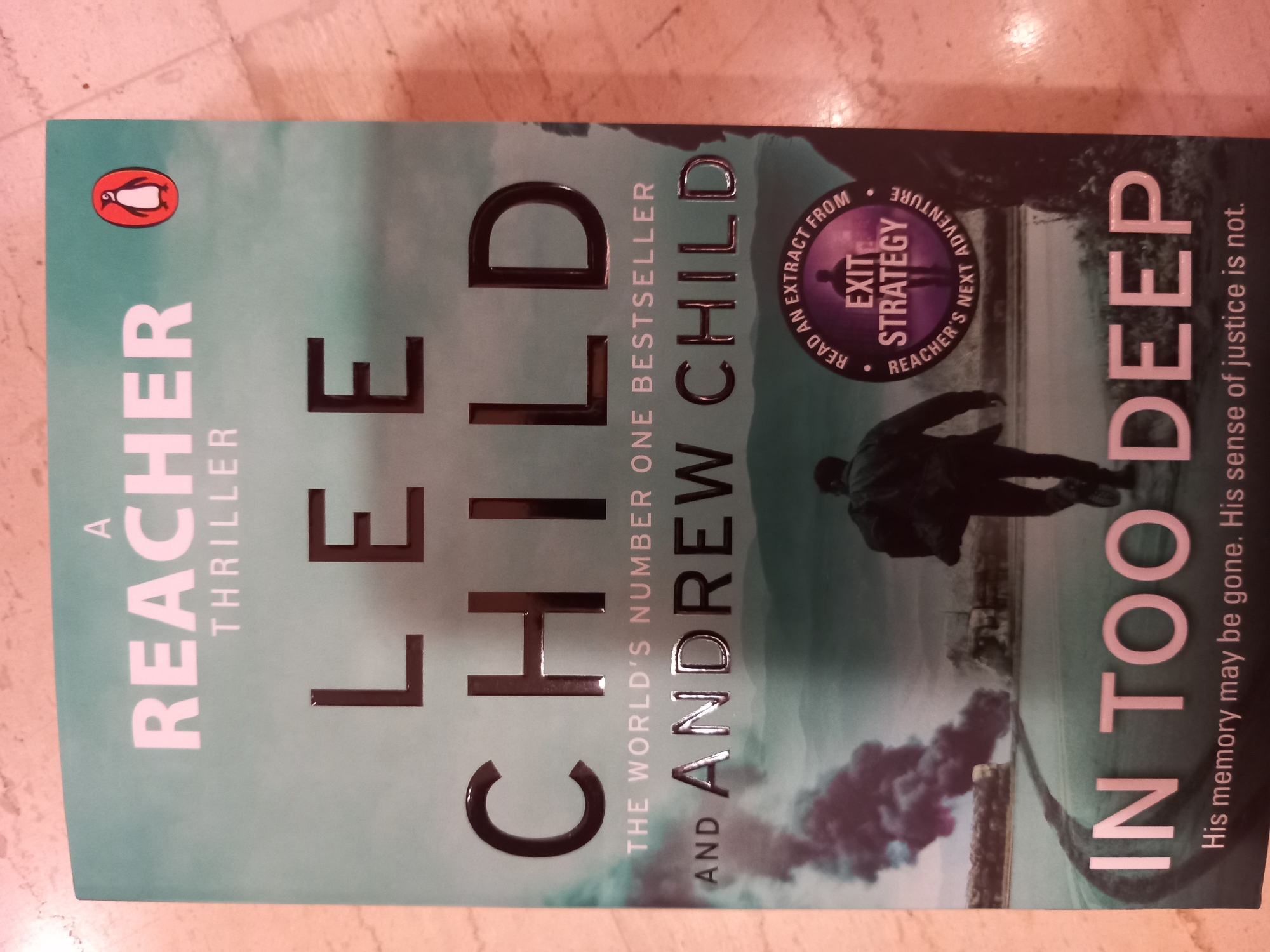 The image is a close-up shot of the cover of a book titled "IN TOO DEEP" by Lee Child and Andrew Child. The book cover features a dark, atmospheric image of a person walking away from the camera on a road with smoke billowing in the background. A circular graphic with the words "READ AN EXTRACT FROM EXIT STRATEGY REACHER'S NEXT ADVENTURE" is superimposed on the image. The title "IN TOO DEEP" is prominently displayed in large, pale pink letters at the bottom of the cover. The book is lying flat on a surface that appears to be a floor with a tiled pattern. The overall lighting suggests an indoor setting with artificial light. There is no discernible city or location information present in the image.