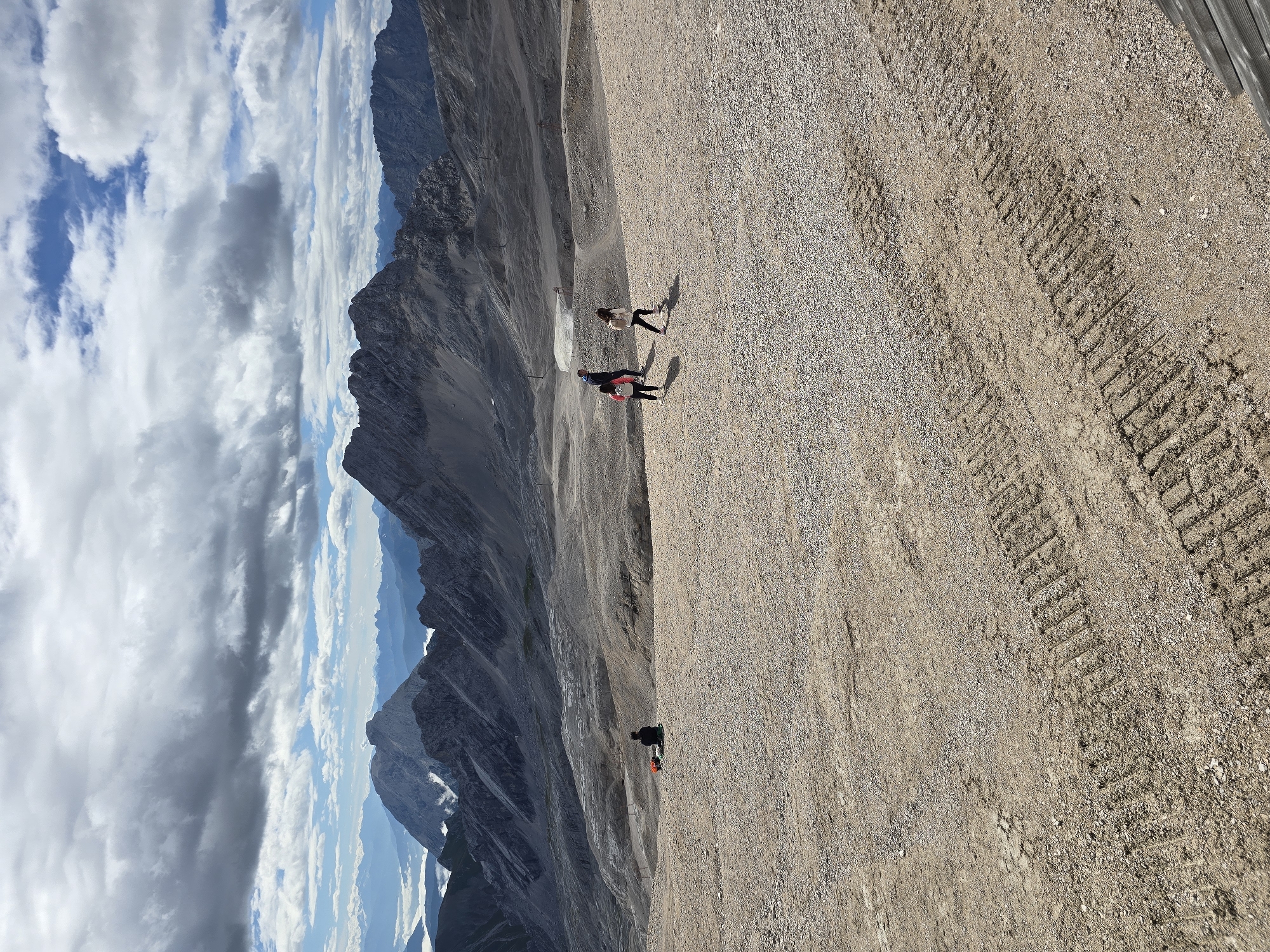 The image shows a wide, barren mountain landscape with a vast expanse of gravel and dirt in the foreground. Several people are visible in the distance. On the left, a lone individual sits with their back to the camera, accompanied by a red backpack and a black bag. Further to the right, a group of three people, two adults and a child, are walking away from the camera. The middle ground features rolling hills of gravel and rock, with a few scattered objects that could be signs or equipment. The background is dominated by imposing, rugged mountain ranges under a dramatic sky filled with dark, heavy clouds interspersed with patches of blue sky. The overall impression is one of a remote, high-altitude environment, likely a mountain peak or plateau in the Alps. This scene is consistent with the summit area of the Zugspitze, Germany's highest mountain, located near Garmisch-Partenkirchen. The tire tracks visible in the foreground suggest recent vehicle activity on the gravel surface.