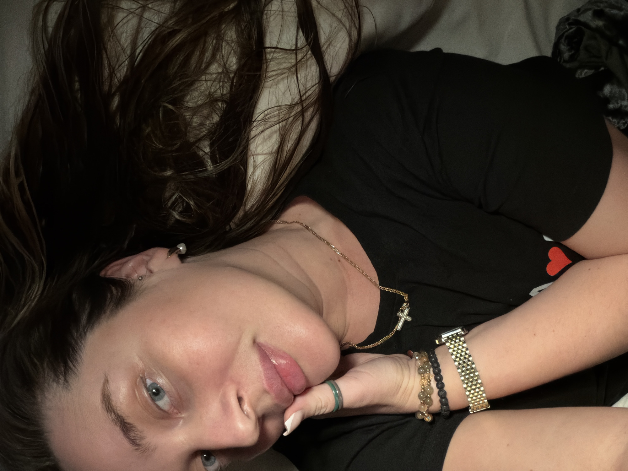 A woman is lying on her side, seemingly on a bed, with a pillow behind her head. Her dark brown hair is spread out around her face and over the pillow. She is wearing a black t-shirt. She is also wearing a gold necklace with a cross pendant. On her wrist, she has several bracelets, including a gold watch. She has blue eyes and fair skin. Her left hand is near her face. Her nails are painted white. No evidence points to the location of Moscow, Russia.