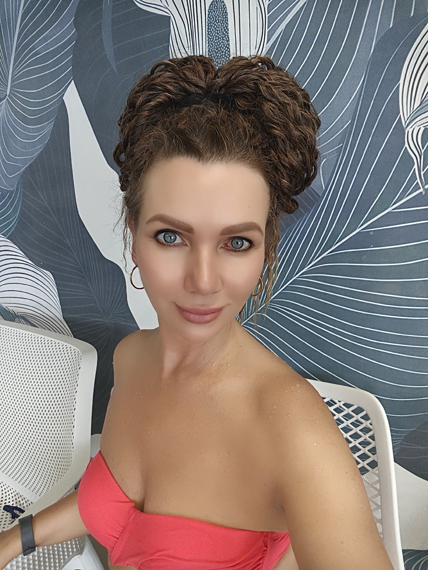 A woman with curly brown hair pulled up in a bun and bright blue eyes is sitting in a chair in front of a wall with a leaf pattern. She is wearing a coral-colored strapless bikini top. Her skin appears to be slightly wet, with small water droplets visible on her shoulders and chest. Her hair is styled in an intricate updo. She is wearing a hoop earring in her left ear. To her left, another white mesh chair is visible. The background features a wall with a repeating pattern of large, stylized leaves in shades of blue and white. The lighting suggests an indoor setting, possibly a spa or sauna. The image is a close-up shot, focusing on the woman's face and upper body.