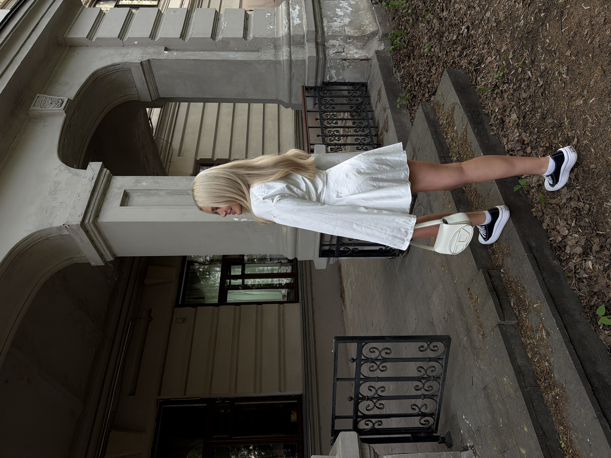 A woman with long blonde hair is standing on the steps of a building, wearing a white dress and black and white sneakers. She is holding a white purse. The building has a grey facade with arched doorways and decorative iron railings. The steps are made of stone and are covered with leaves. The location is Kazan, Russia.