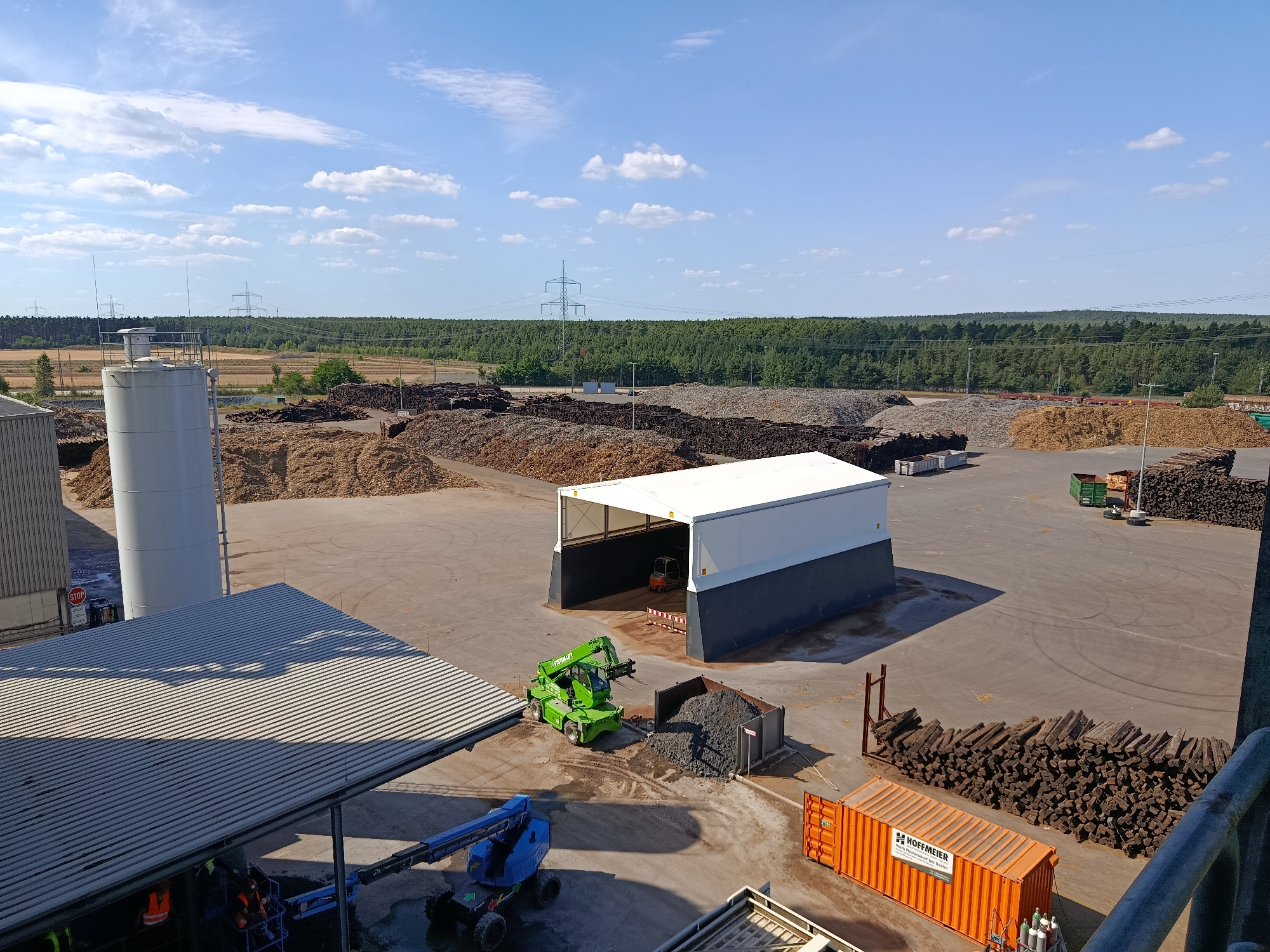 This is an aerial view of an industrial site, possibly a biomass processing facility, in Baruth/Mark, Germany. The scene is bathed in the bright, clear light of a sunny day, with scattered white clouds against a vibrant blue sky.
Dominating the foreground is a large, corrugated metal roofed structure, partially shaded. Underneath it, two individuals in high-visibility vests are working on a blue articulated boom lift. Adjacent to this structure, a green telehandler with a fork attachment is parked near a large metal skip filled with dark, granular material. A small orange forklift is visible inside a white and dark grey open-sided industrial building in the middle ground.
The yard is paved and shows tire marks. Large piles of wood chips and other biomass materials are stacked throughout the site, some appearing freshly processed and others weathered. In the background, a dense forest forms a green horizon, with power lines and transmission towers stretching across the landscape.
On the right side of the image, a bright orange shipping container with the text "HOFFMEIER" and "www.hoffmeier-berlin.de" is parked next to a neatly stacked pile of dark, cuboid-shaped material, possibly peat or compressed biomass. Another green skip is visible further in the distance.
The overall impression is one of an active industrial operation under fair weather conditions, likely during daylight hours. The presence of various machinery and the extensive piles of processed material suggest ongoing work related to resource management or energy production.